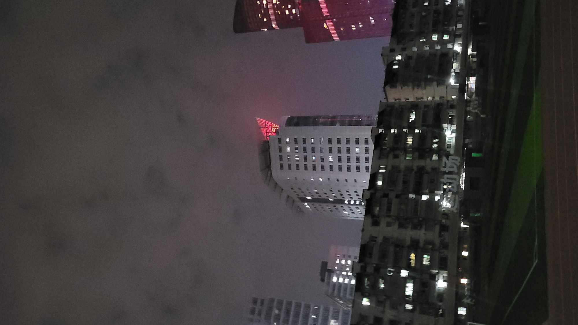 At night, a view upward captures buildings in Yuzhong District, China. A tall white building with a red illuminated crown stands out. Other buildings with lit windows surround it. Fog or clouds obscure the sky. A green and white sign hangs on one building, partially visible. The picture is rotated.