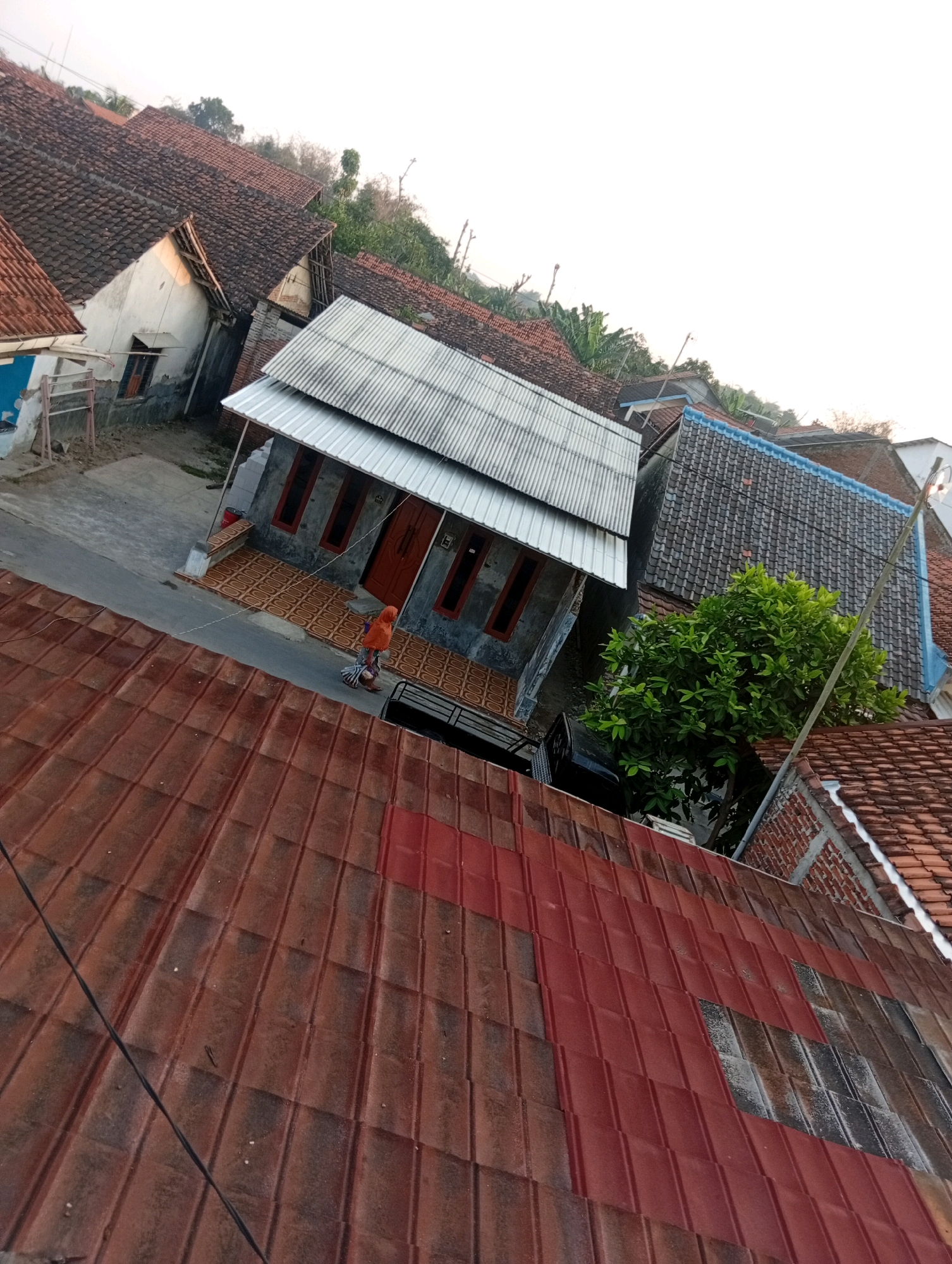 In the city of Kebandingan, Indonesia, this high-angle, slightly tilted photograph captures a residential scene. In the foreground, a reddish-brown tiled roof dominates, showing signs of weathering. Beyond it, a narrow paved street runs diagonally. On the street, a child wearing an orange headscarf and a grey-patterned garment is riding a scooter. The child appears to be moving away from the viewer. Adjacent to the street is a small, gray house with a metal roof, featuring three prominent windows with red frames and a red door. The house is adorned with patterned tiles on the porch. To the right of this house, a lush green tree stands out against the backdrop of other houses with tiled roofs, some blue-edged. The overall environment suggests a typical Indonesian neighborhood with close-knit housing and verdant vegetation. The sky is bright and appears to be late afternoon or early morning, indicated by the soft light.