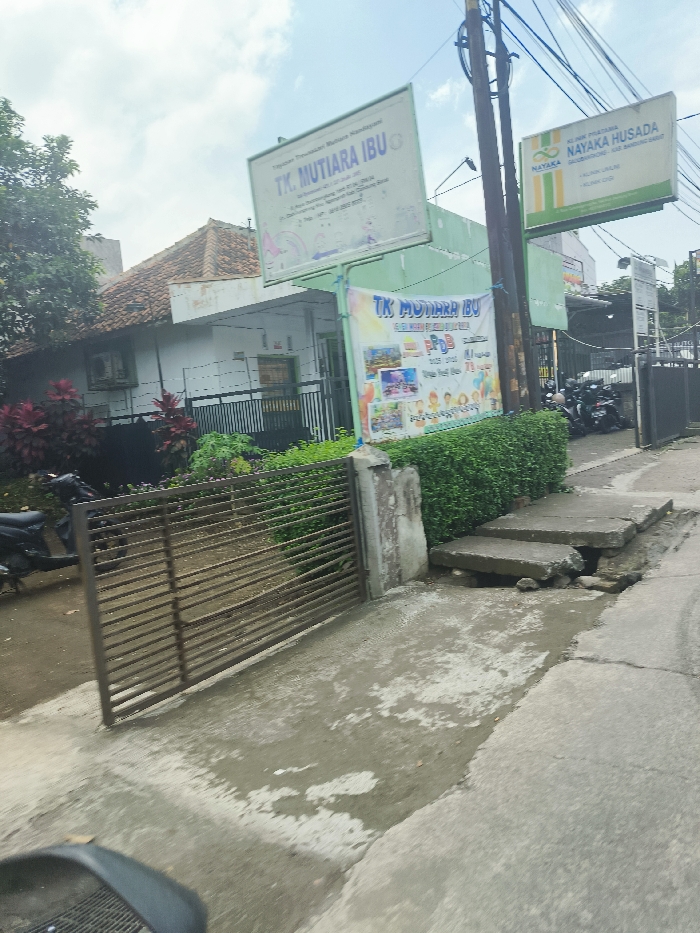 The image shows a street in Ngamprah, Indonesia, with a building on the left side, labeled "TK MUTIARA IBU," a kindergarten. In front of the building is a metal gate, and a black motorcycle is parked next to it. There is a small garden area with green shrubs and a few potted plants with red flowers. The street is paved with concrete and appears to be somewhat weathered. There is a set of concrete steps that appear to be broken on the right side of the street. Further down the street, on the right side, there are more buildings and some additional motorcycles are parked. There are two signs visible; one on the left side of the building, labeled "TK MUTIARA IBU," and another on the right side, labeled "YAYASAN NAYAKA HUSADA." The sky is overcast with white clouds.