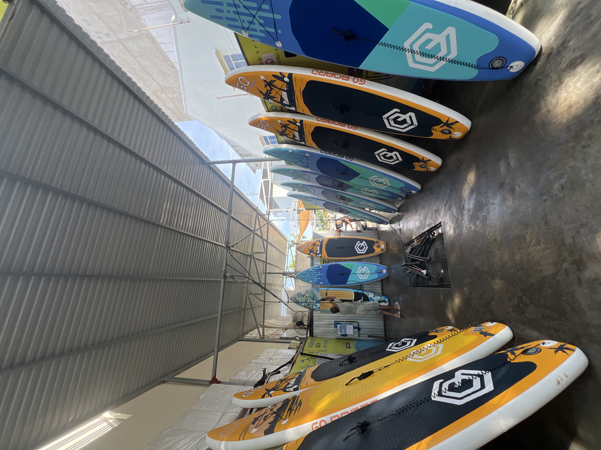 In Bac Nha Trang Ward, Vietnam, a covered outdoor storage area is filled with numerous stand-up paddleboards. The boards are arranged in rows, leaning against a corrugated metal wall. The majority of the paddleboards are colorful, with designs featuring palm trees and the "GO BOARD" logo. One man is visible near the center of the image, appearing to interact with one of the paddleboards. The ground is wet concrete, suggesting a recent wash or damp conditions. In the background, a building and the sky are visible.