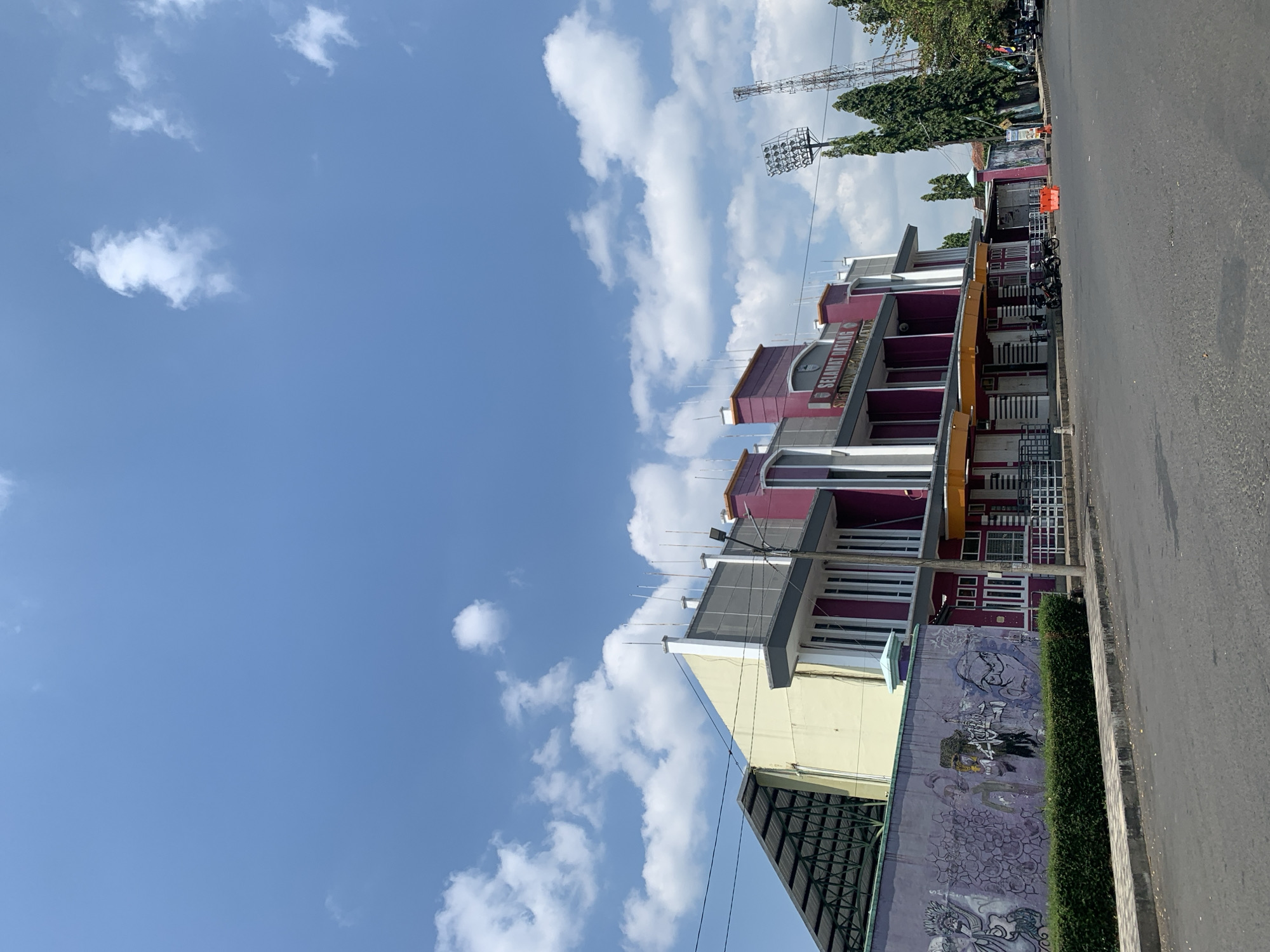 The image captures a bright, sunny day in Ciamis, Indonesia, with a clear blue sky dotted with white, fluffy clouds. In the foreground, a paved road runs horizontally across the frame, with a sidewalk bordering it. A low, manicured hedge runs along the edge of the sidewalk.
Behind the hedge, a long, vibrant purple mural adorns a wall. The mural features various graphic illustrations and abstract figures in shades of black, white, and purple.
The main subject of the image is a building with a distinctive architectural style. The building features a prominent section painted in a deep magenta color, with white vertical window frames. To the left of this section, another part of the building is painted in a pale yellow, with a sloped, dark-colored roof structure extending outwards. The building has a modern, functional design with awnings over the lower entrances. A sign above one of the entrances reads "SELAMAT DATANG STADION GALUH CIAMIS," indicating that this is the welcome entrance to the Galuh Stadium in Ciamis.
To the right of the magenta section of the building, a tall, slender cell tower stands prominently against the sky. Further to the right, a stadium light tower with multiple bright lights is visible, suggesting the presence of a sports facility. Several trees, some tall and slender and others with fuller canopies, are interspersed with the structures.
In front of the building, a few parked motorcycles and an orange construction barrier are visible on the road, indicating ongoing activity or preparation. The overall scene portrays an urban environment, likely on the outskirts or within a sports complex in Ciamis.