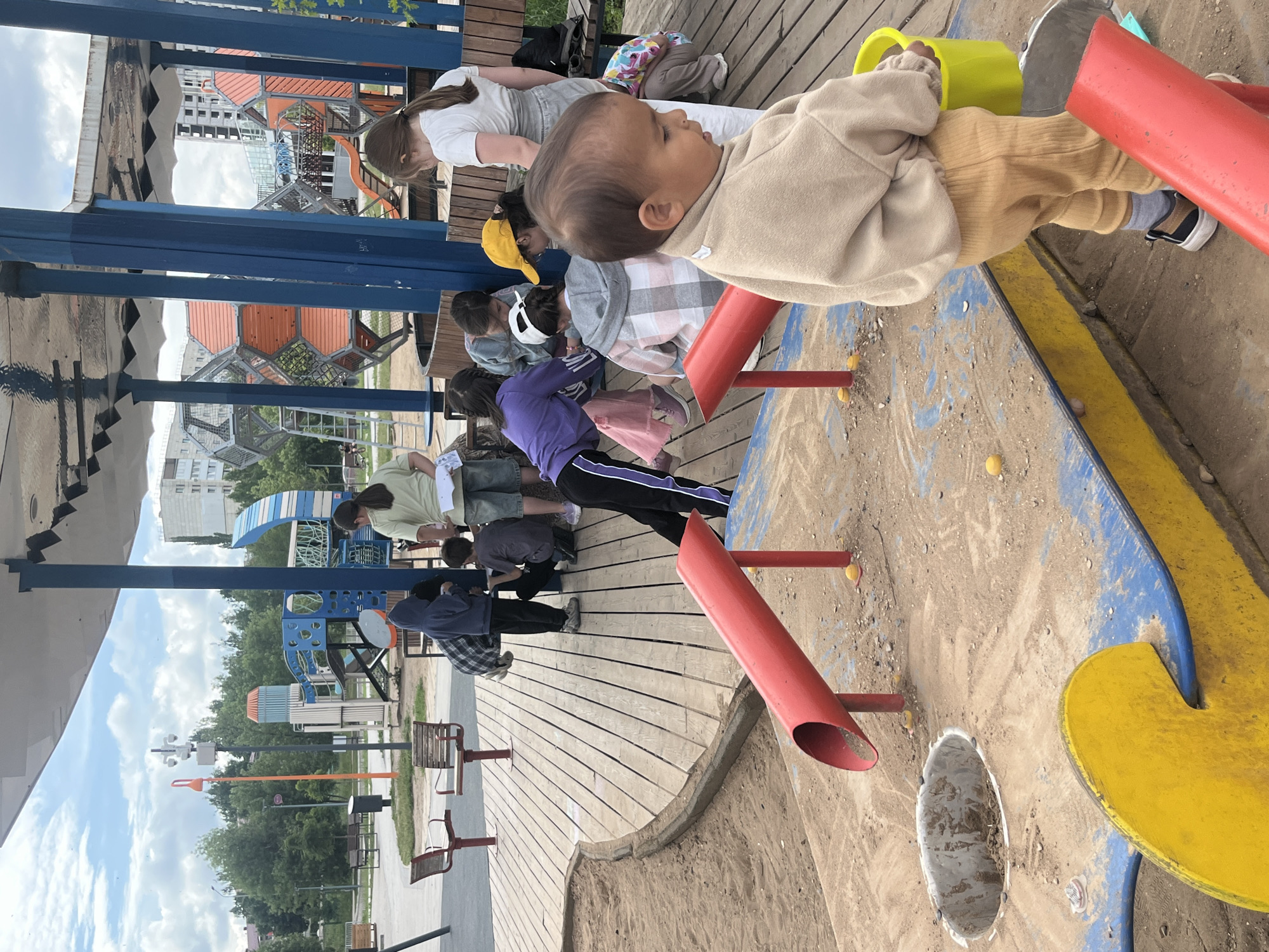 The image depicts a group of children and an adult in a playground area, likely located in the city of Oktyabrsky, Russia. In the foreground, a young child, appearing to be a toddler, is dressed in a tan sweatshirt and tan ribbed pants, with a yellow bucket in their right hand. They are standing on a sand-covered surface with blue and yellow playground structures. Several other children are gathered around a wooden deck in the background, engaged in various activities. Some appear to be looking down at papers or objects. The playground features red pipes, a circular opening in the sand, and wooden benches. In the middle ground, there are larger playground structures with orange and blue geometric shapes and slides. The background reveals residential buildings and trees under a cloudy sky. The overall scene suggests a casual, outdoor gathering or activity in a public space.
