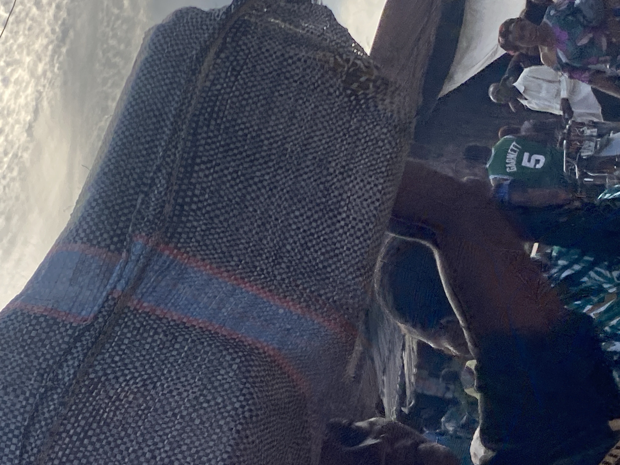 A close-up, tilted image shows a crowd, possibly at an event in Igbara, Nigeria. The subject is partially obscured by a large, textured cloth, dark in color, with a blue and red stripe across it. Behind the cloth, a person wearing a green jersey with the number "5" and the name "GARNETT" on the back is visible, along with other people in the background. The sky appears cloudy. The crowd suggests a public gathering, but the exact nature of the event is unclear.