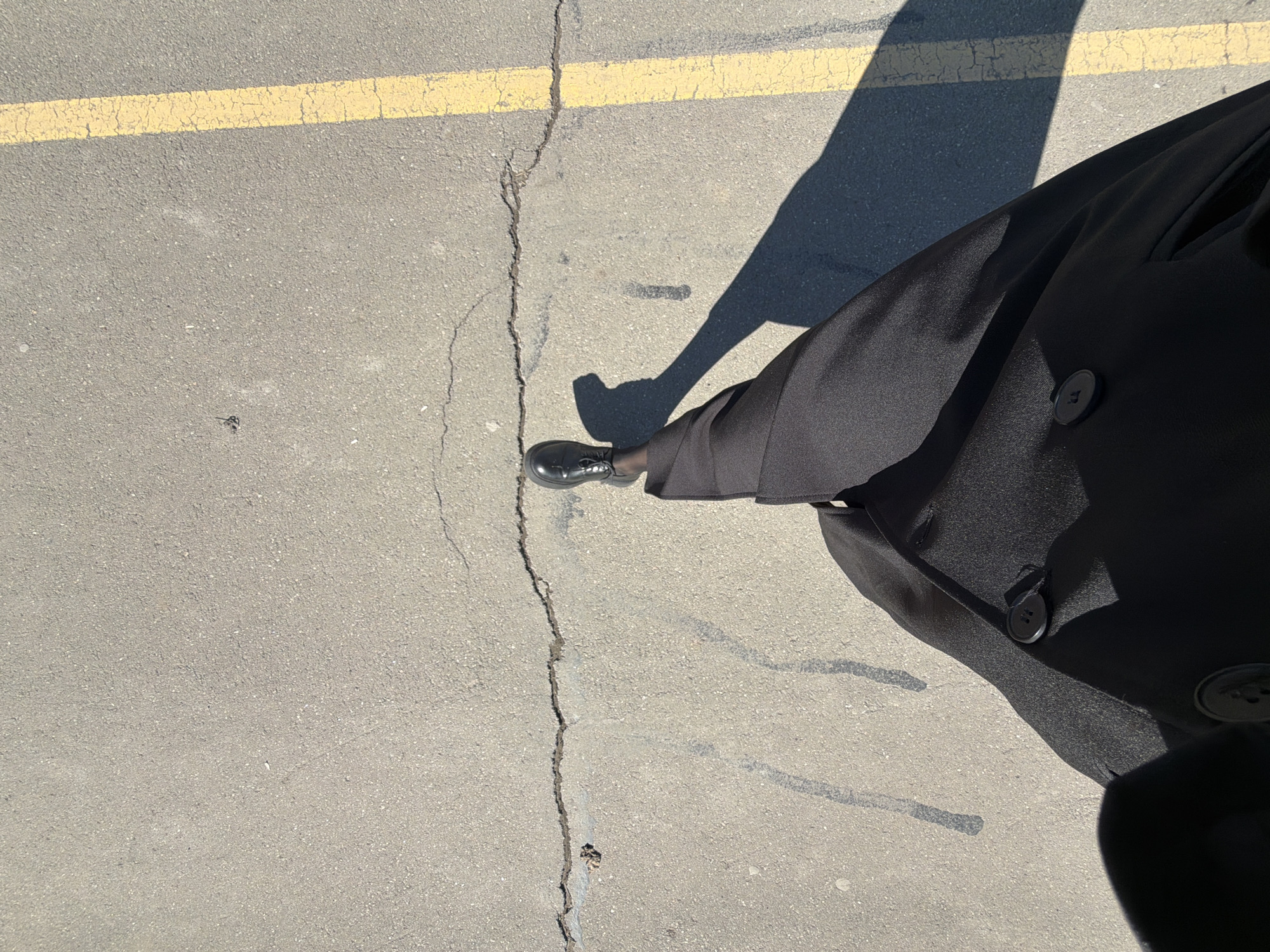 An aerial shot depicts a portion of asphalt pavement marked with a yellow line at the top left. A crack runs diagonally across the frame from the top left to the bottom center. The lower right quadrant features the lower portion of a person clad in a dark, buttoned coat and black leather shoe. The shadow of the person is visible, cast diagonally from the top right to the lower middle. The scene is outdoors, illuminated by natural light. The location is not possible to determine.