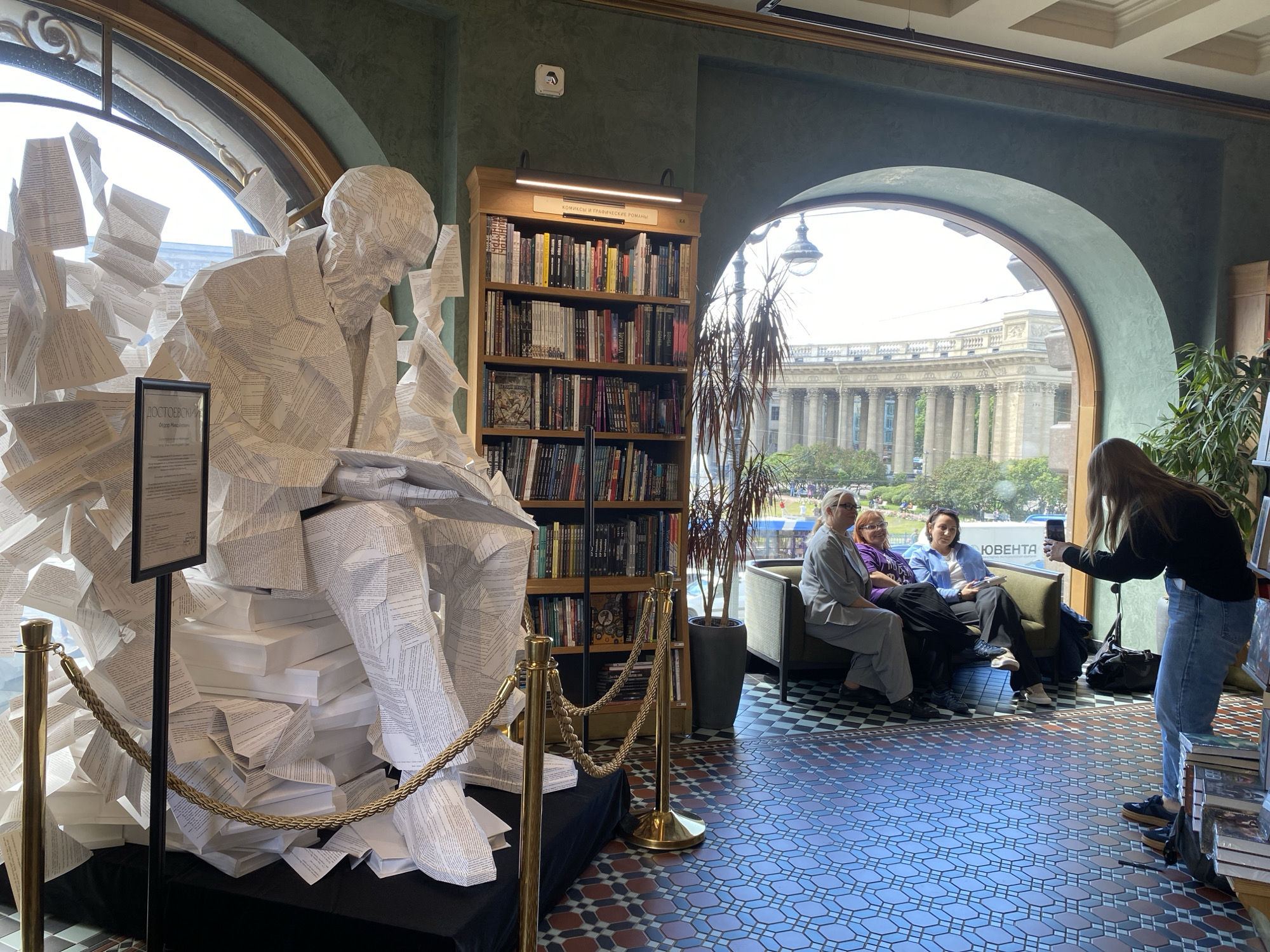 An interior view of the Dom Knigi (House of Books) bookstore in Saint Petersburg, Russia, showcases a prominent sculpture of Fyodor Dostoevsky made from folded book pages. The sculpture is positioned near a large arched window, with stacks of white books forming its base. In the background, a tall bookshelf filled with books is visible, as are several patrons seated on a couch near another large window offering a view of Kazan Cathedral. A woman is standing, taking a photograph of the seated individuals with her smartphone. The floor features a distinctive patterned tile design. The lighting is natural, coming from the windows, and supplemented by interior lighting.