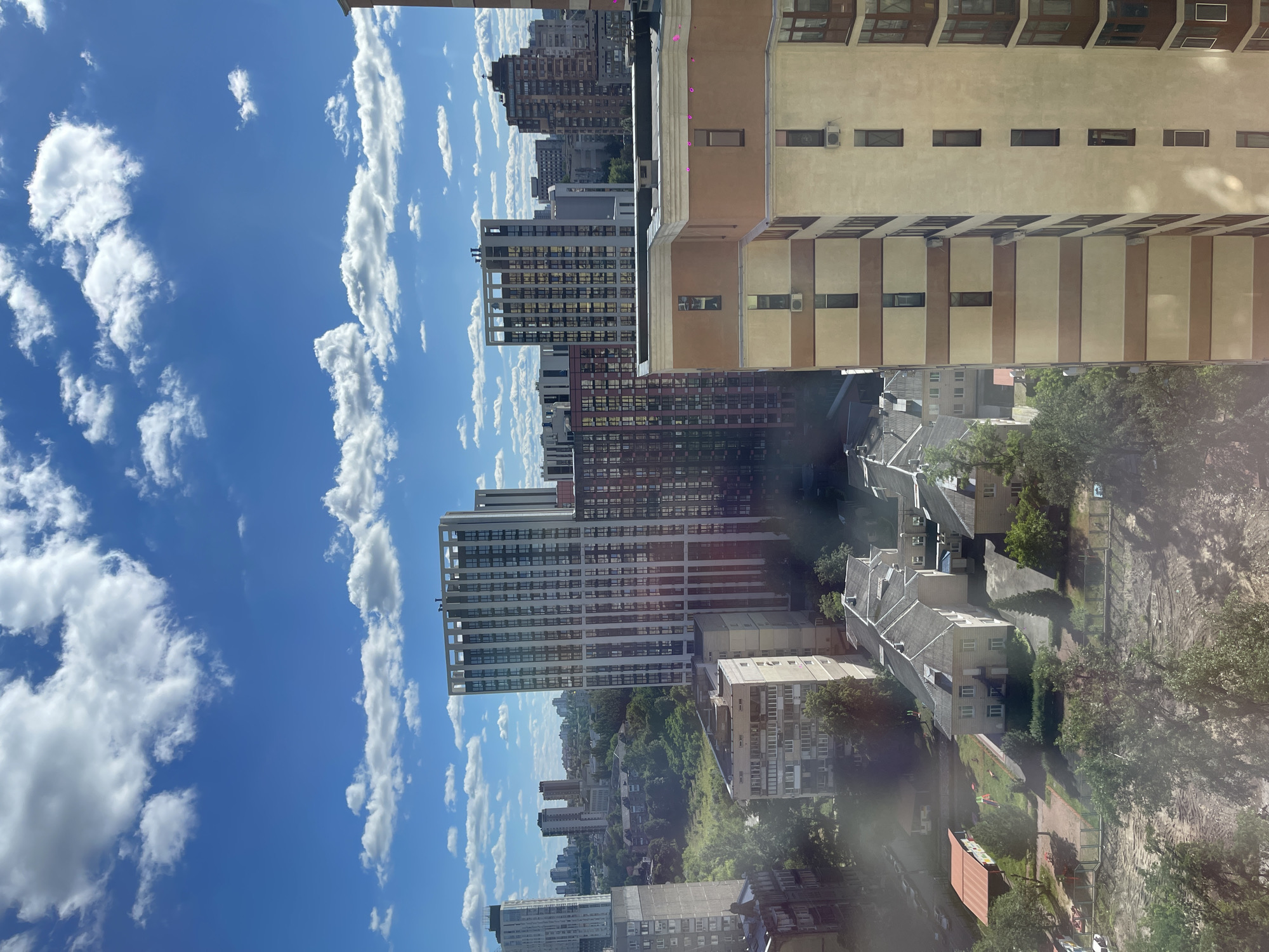 The image shows a cityscape with multiple buildings under a bright blue sky with white clouds. In the foreground, a tall, light-colored apartment building with brown horizontal stripes and many windows dominates the right side of the frame. To the left of this building, a row of smaller residential buildings with grey roofs are visible. Further in the background, several taller, modern apartment buildings with varied architectural designs and colors are spread across the urban landscape, interspersed with green trees and vegetation. The scene appears to be a residential area in Kyiv, Ukraine, likely captured from an elevated vantage point. There are no people or vehicles clearly visible in the image. The overall impression is that of a sunny day in a densely populated urban environment.
