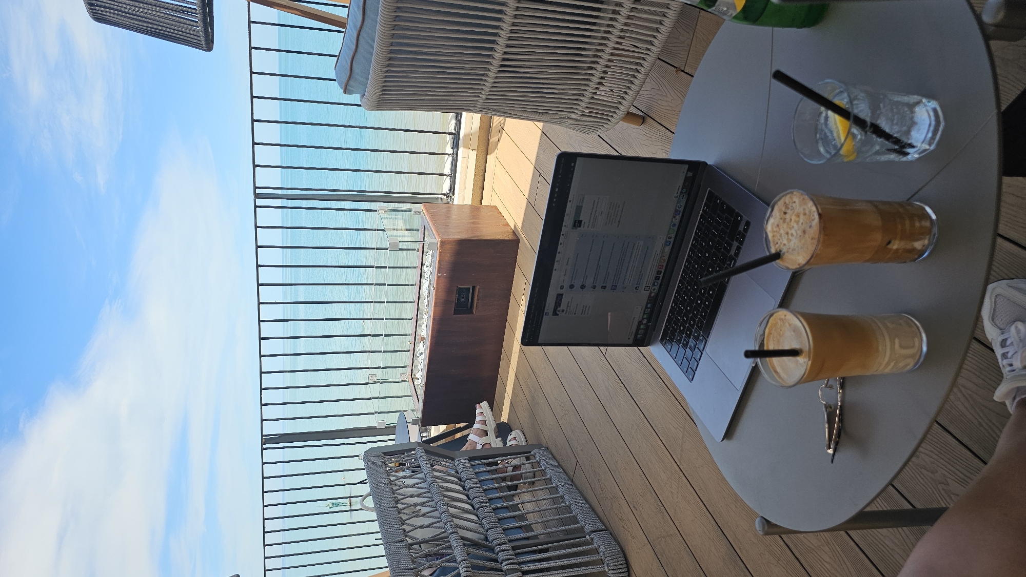 The media file depicts an outdoor scene, likely a balcony or terrace overlooking the sea. The viewpoint is from a seated position, looking down and across. The primary subjects are a laptop, two glasses of iced coffee with straws, and a glass of water with a lemon slice and a straw. The laptop is open and appears to be in use, displaying text and possibly a website or application. One of the iced coffees is closer to the camera, while the other is slightly further back. The glass of water is to the left of the coffees.
The setting is a modern outdoor area with wooden decking. A grey circular table holds the drinks and the laptop. In the background, there is a black metal railing separating the terrace from the view beyond. Through the railing, the turquoise sea is visible under a bright blue sky with scattered white clouds. A dark, rectangular object, possibly a planter or a fire pit, is also present in the mid-ground. To the left, a portion of a wicker chair and a cushioned seat can be seen, suggesting a comfortable seating arrangement. To the right, a person's foot wearing a white sneaker is visible at the edge of the frame, implying someone is present and using the laptop.
The scene suggests a relaxed and productive atmosphere. The bright daylight and clear sky indicate it's daytime, likely a sunny day. The presence of iced drinks and the sea view hint at a warm weather location. The context provided places the location as Varna, Bulgaria, which aligns with a seaside setting. There is no visible text in the image that provides specific information. The overall mood is calm and inviting, suitable for working or enjoying a beverage outdoors.