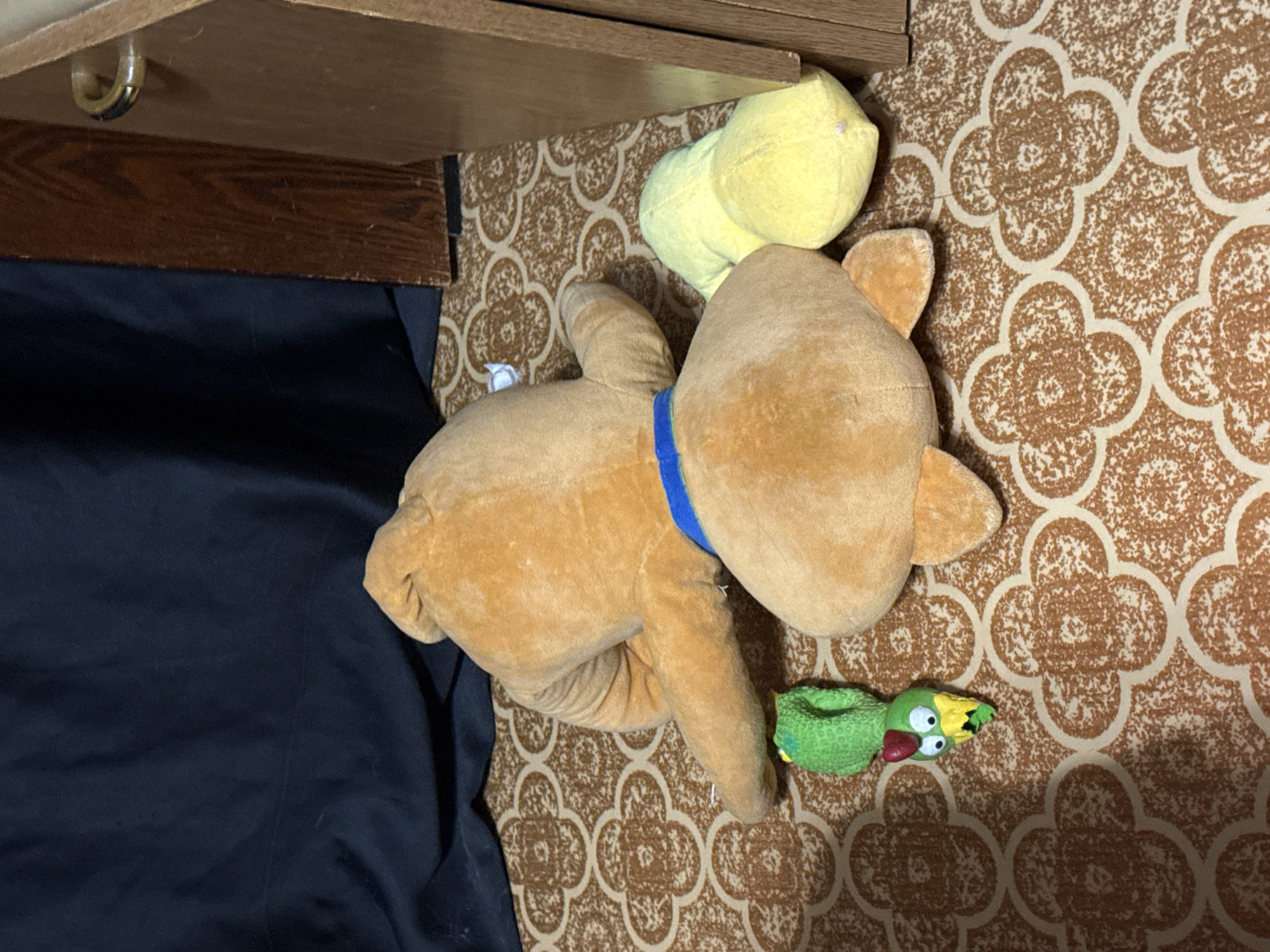 In a room with patterned carpet, a large tan stuffed dog with a blue collar lies on its side. To its left is a small green stuffed snake with red eyes and a red heart on its head. To the right of the dog is a yellow stuffed toy. A dark blue curtain is visible in the background on the left, and a wooden cabinet door with a brass handle is on the right. The image does not contain any information that would allow for its location to be identified in Khimki, Russia.