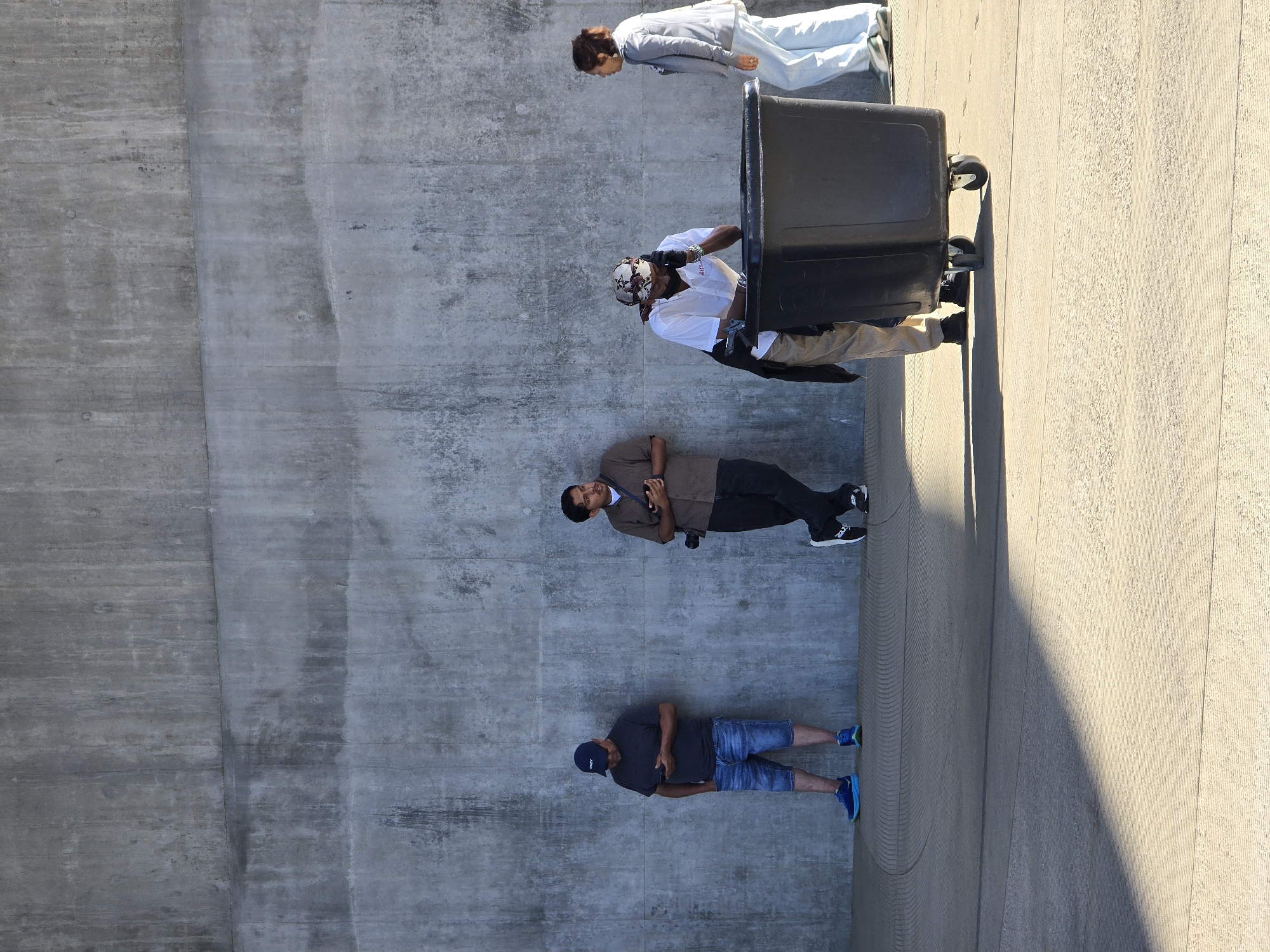 Four individuals are depicted leaning against a textured concrete wall in an outdoor urban environment. The wall is the dominant background element, exhibiting a rough, industrial aesthetic with horizontal lines and variations in tone. In the foreground, a textured concrete ground plane is illuminated by harsh sunlight, creating a distinct shadow on the left side of the frame.
From left to right, the first individual is a person wearing a dark t-shirt, denim shorts, and a blue baseball cap. They are standing with one hand in their pocket and looking down at a mobile phone. Adjacent to them, a second individual, dressed in a brown t-shirt and dark pants, holds a camera around their neck and appears to be looking to their right.
Further to the right, a third individual, wearing a white shirt, patterned bandana, and dark pants, is bent over a large black rolling trash bin, holding a phone to their ear. A dark jacket is slung over their shoulder. To the far right, a fourth individual, in a light-colored jacket and light-colored pants, is walking away from the trash bin, looking towards the left.
The overall scene suggests a moment of pause or waiting, with individuals positioned along the wall. The environment appears to be public space, possibly near a walkway or park. The specific location in Los Angeles, United States of America, cannot be definitively determined from the visual information alone.