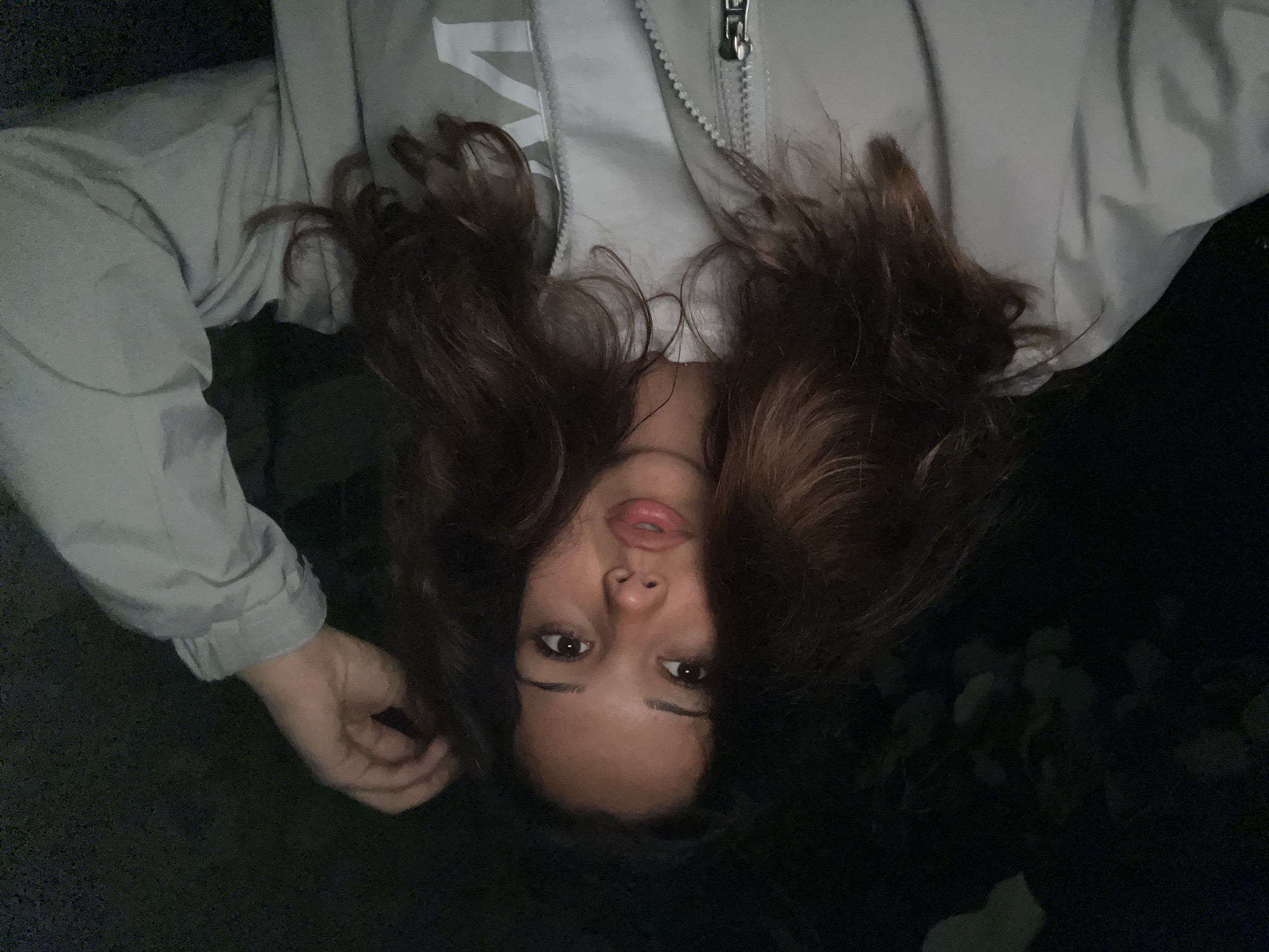 A young woman is captured in a selfie, taken outdoors in what appears to be nighttime. She is wearing a light-colored zippered jacket over a white t-shirt. Her long, dark, wavy hair is partially covering her face, and her right hand is raised, touching her hair. Her lips are slightly parted, and she is looking towards the camera. The background is dark, with faint hints of foliage suggesting a natural setting, possibly a park or garden. There are no discernible landmarks or features that would indicate a specific location within Zlatograd, Bulgaria.