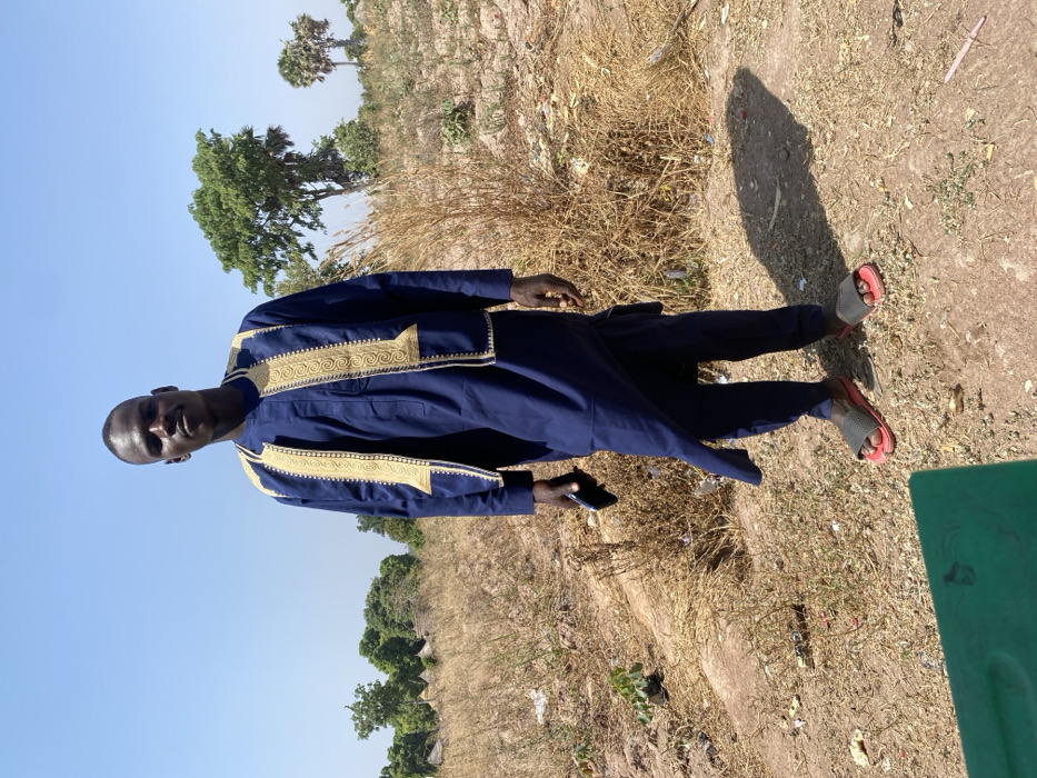 A dark-skinned male, approximately in his twenties to thirties, stands facing forward, looking directly at the camera. He wears a dark blue traditional garment consisting of a tunic and trousers, with a gold-embroidered vest worn over the tunic. He holds a dark-colored smartphone in his left hand, down by his side. His feet are clad in black and red slide sandals.
The individual is positioned outdoors on dry, light brown soil with sparse, straw-colored vegetation and scattered debris, including what appears to be plastic. In the background to the left, several small, round, earth-colored structures with conical roofs are visible amidst trees, suggesting a rural settlement. To the right, mature green trees and two distinct palm trees stand against a clear, light blue sky. Strong overhead sunlight creates a distinct shadow of the individual on the ground to his right. A partial green object is visible in the lower left foreground. The scene is located in Zaki Biyem, Nigeria.