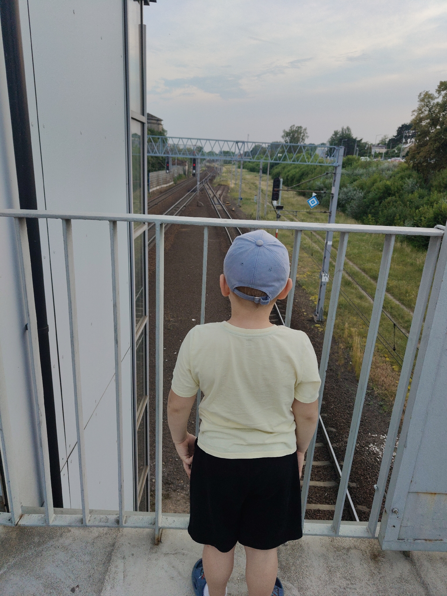 A young boy, facing away from the camera, stands on a pedestrian bridge overlooking a set of train tracks. He is wearing a light yellow t-shirt, black shorts, and a light blue baseball cap. The bridge has a light gray metal railing. In the distance, the train tracks curve to the left, with a railway signal displaying a red light. Overhead, a metal structure spans the tracks, likely part of the railway infrastructure. The scene is set during the day, with an overcast sky. Green trees and vegetation line the sides of the tracks. The boy appears to be observing the train tracks, possibly waiting for a train. The location is identified as Nowy Dwór Mazowiecki, Poland.