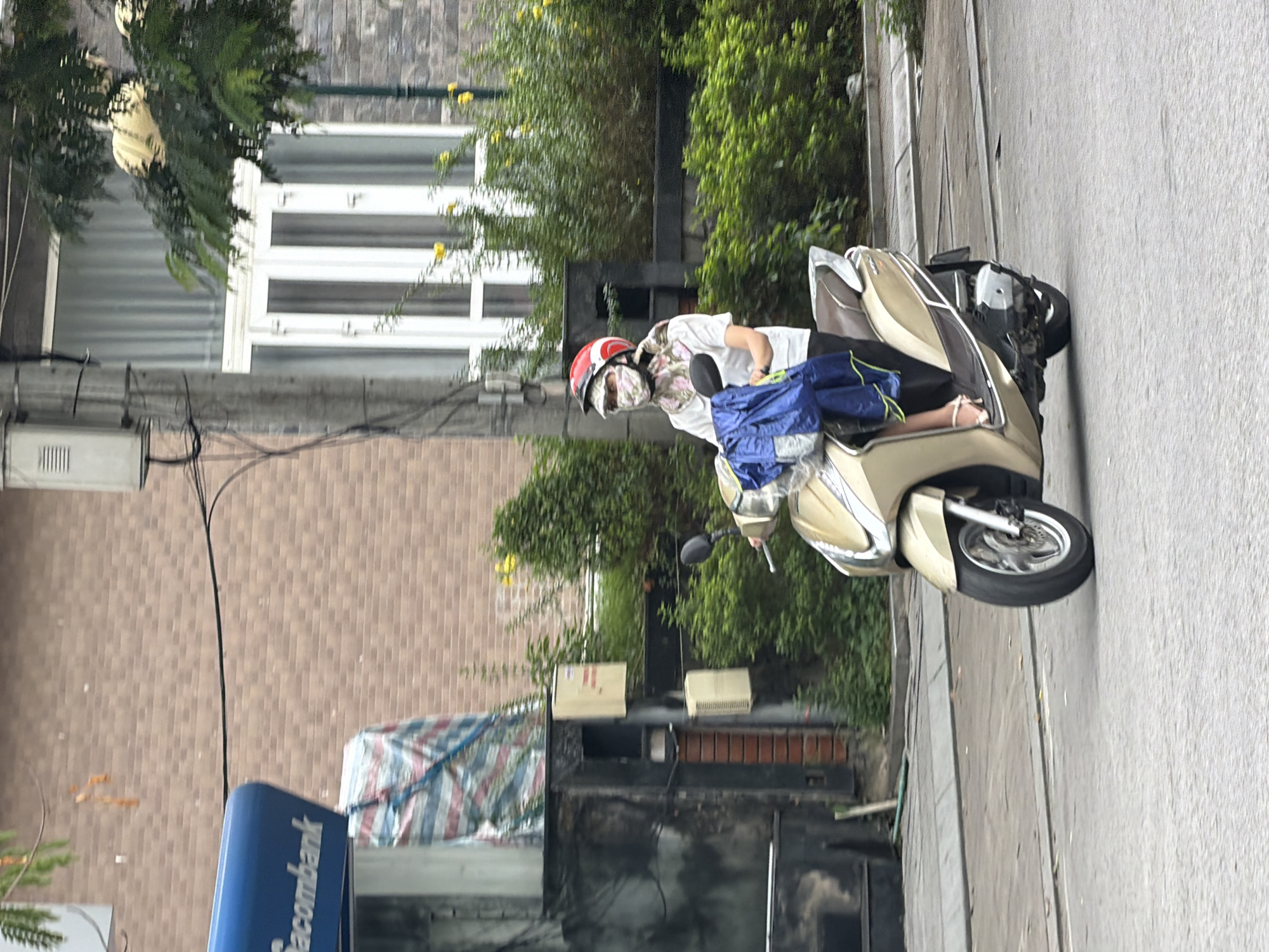 A woman wearing a white shirt, black pants, a floral headscarf, and a red helmet rides a gold scooter on a street in Thuận Hóa Ward, Vietnam. She is holding a blue bag on her lap. The street is paved, and there is a sidewalk to the left of the road. In the background, there is a brick building with a white-framed window and lush green trees. A blue sign with the word "sacombank" is visible on the left side of the image.