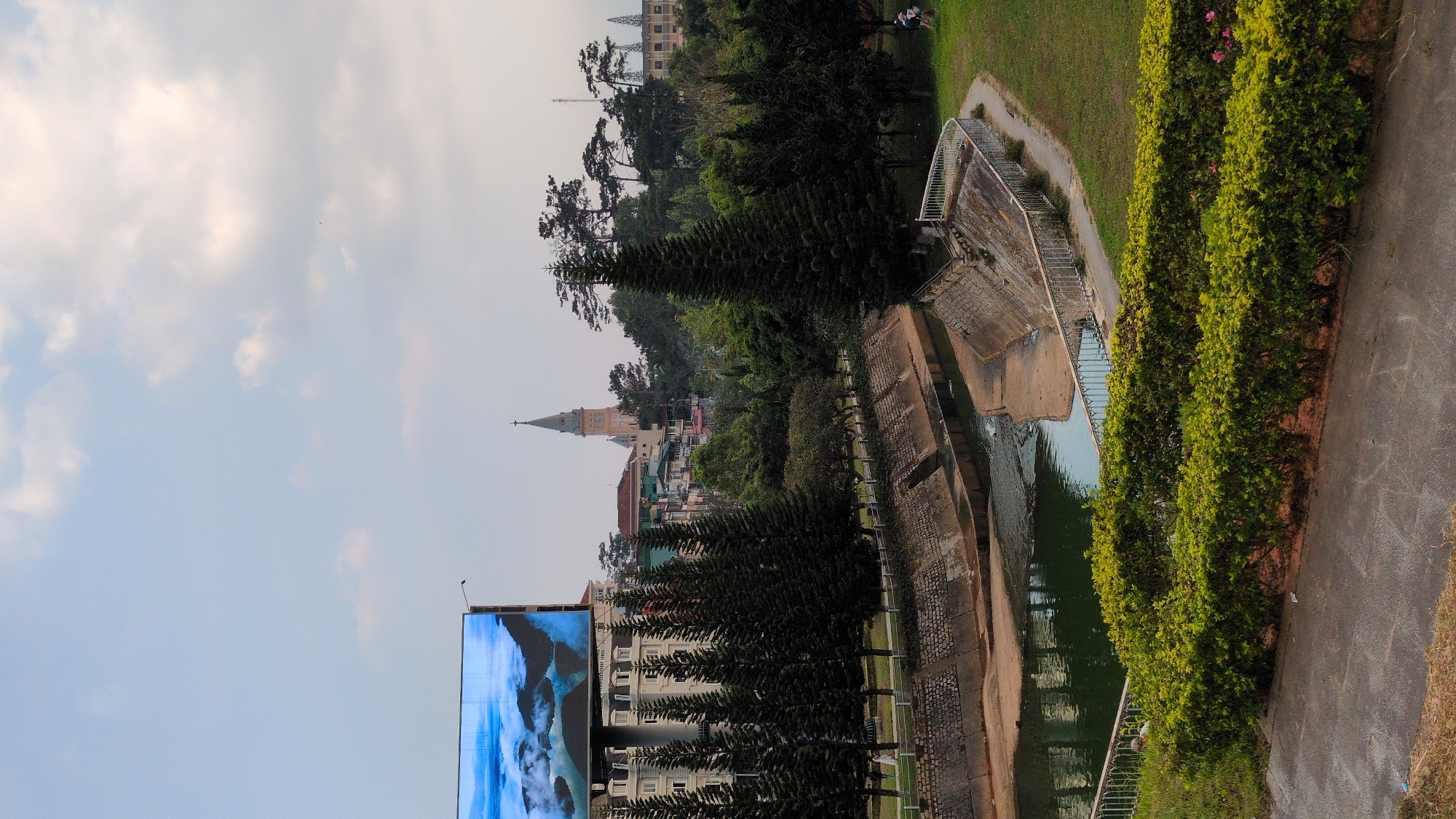 The image shows a view of Da Lat, Vietnam. A large LED screen is visible on the left side. In the distance, a church spire is seen amidst a cityscape of buildings and trees. A water canal runs through the landscape, lined with manicured bushes and a paved pathway. A few people are visible on a grassy area. The sky is partly cloudy.