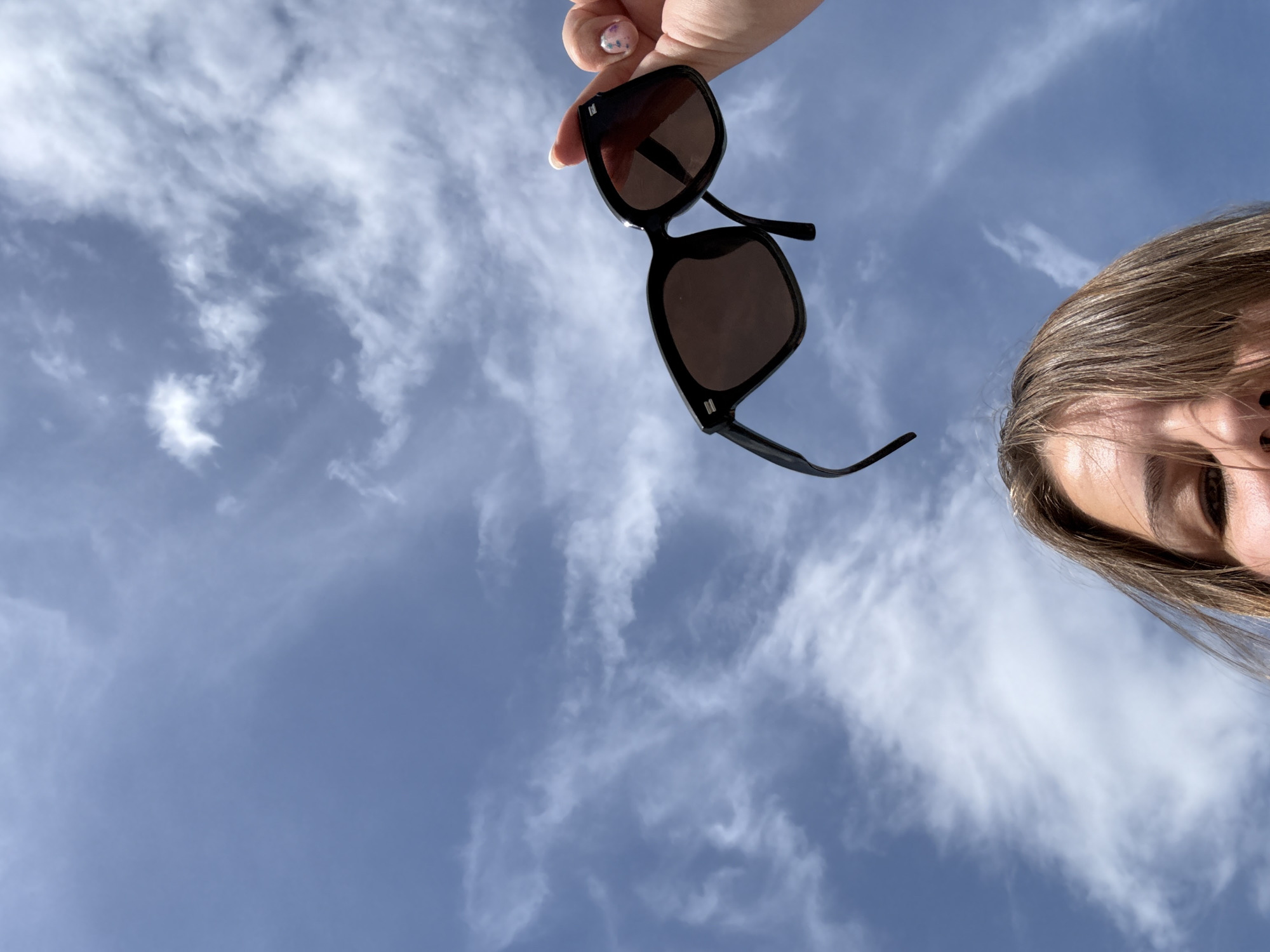 A low-angle shot reveals a person holding a pair of black sunglasses against a backdrop of a blue sky filled with white clouds. The person's right hand is visible holding the sunglasses by the frame. The sunglasses have dark lenses. A portion of the person's face, specifically the forehead, eye, and some hair, is visible on the right side of the frame. The setting appears to be outdoors. The city and country cannot be confirmed with the provided information.