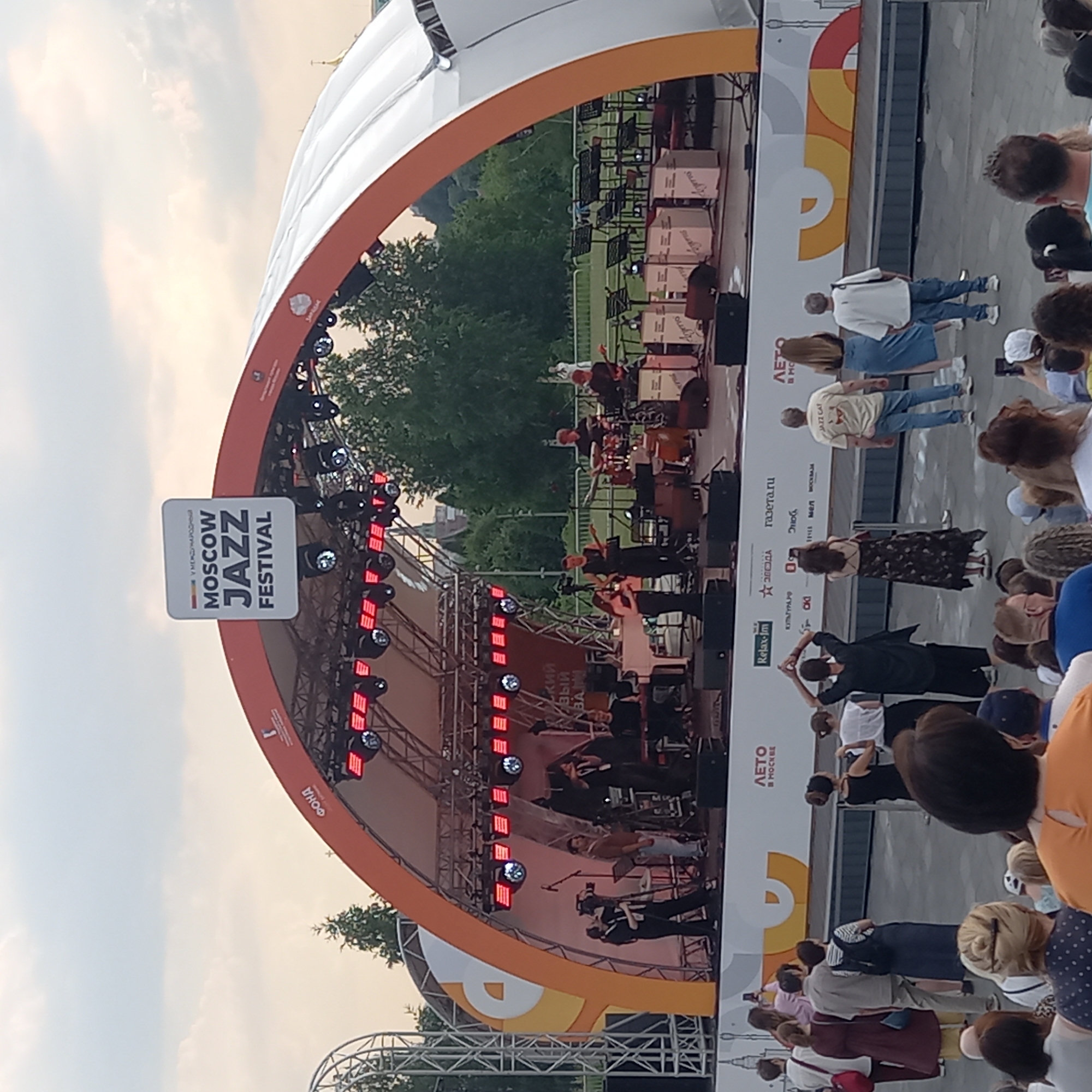 The image shows an outdoor concert stage set up for the Moscow Jazz Festival. A band is performing on stage, consisting of a female singer, a male guitarist, a keyboardist, a drummer, and another guitarist. The stage is adorned with red lights and a large banner at the top reads "Moscow Jazz Festival." In front of the stage, a crowd of people are gathered to watch the performance. The background shows trees and a cloudy sky. The location of the event is stated as Путрышкаўскі сельскі Савет, Belarus.