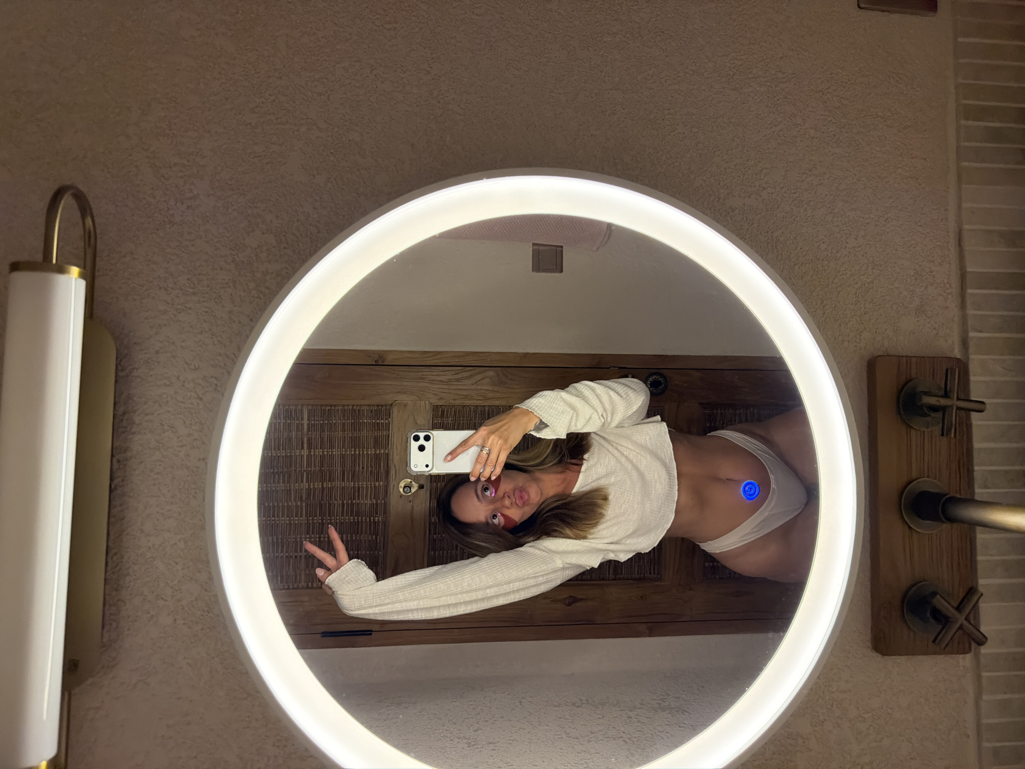 A young Caucasian woman with brown hair is taking a selfie in a circular illuminated mirror in a bathroom. She is wearing a white crop top and white underwear, and has pink under-eye patches on. Her left arm is raised, and she is puckering her lips. The mirror reflects a wooden door and a textured wall. There is a faucet visible below the mirror. The location is a bathroom in Mengwi, Indonesia.