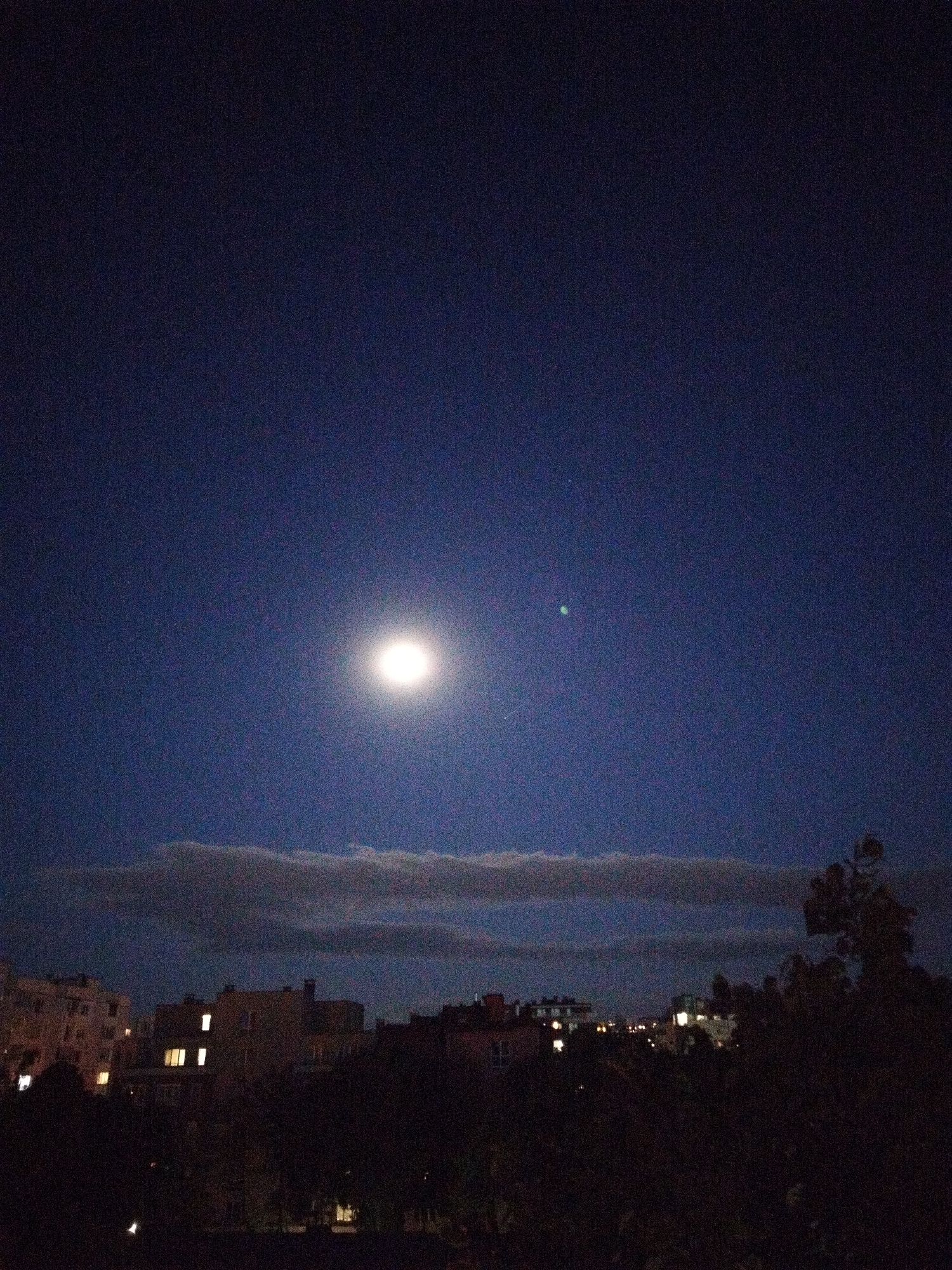 A night scene shows a full moon centered in the dark blue sky above Sofia, Bulgaria. Below the moon, a horizontal band of clouds stretches across the middle of the frame. Dark silhouettes of buildings and trees fill the lower portion of the image. Some windows are illuminated in the buildings. The foreground is mostly dark.