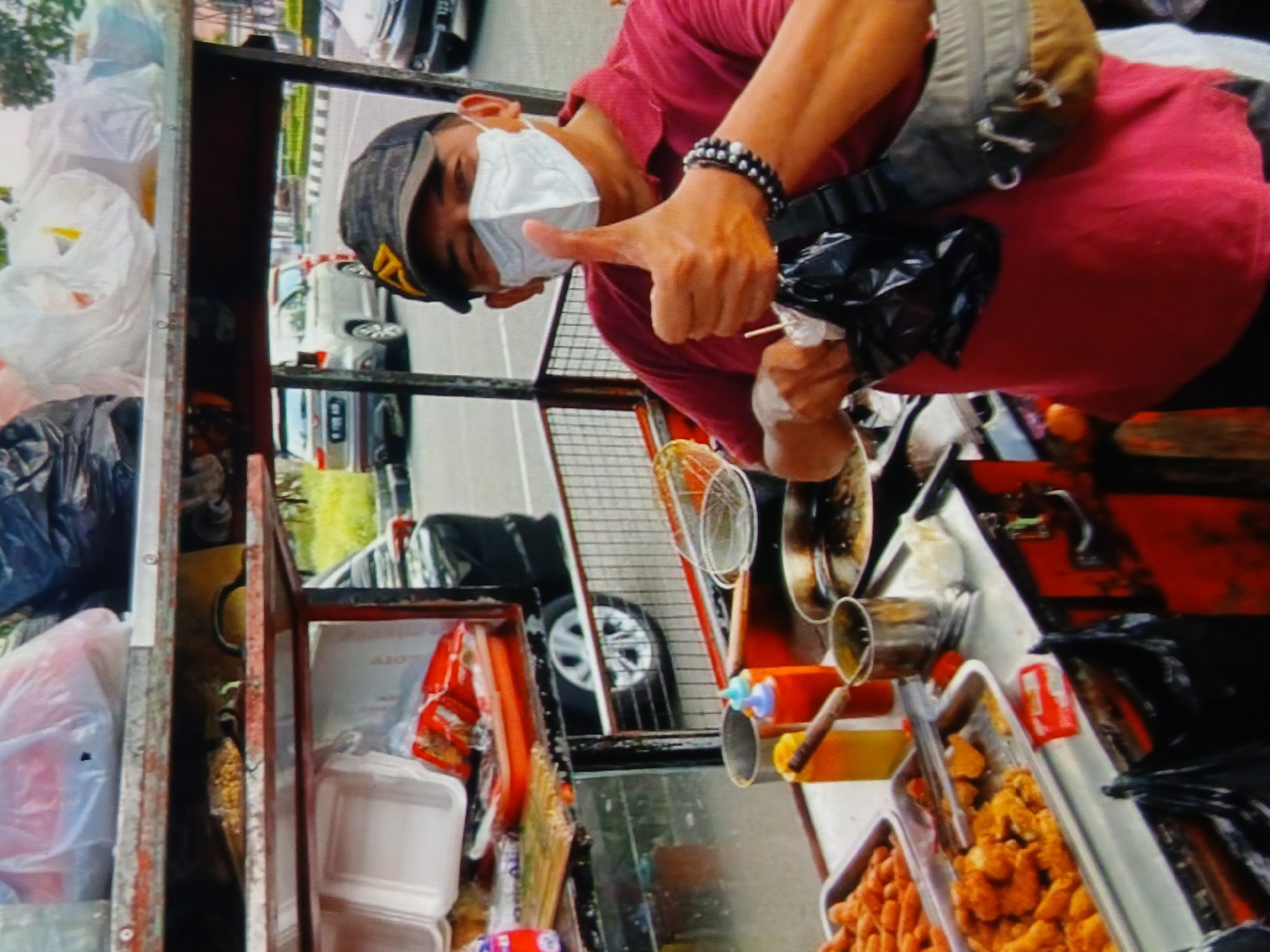 In the city of Setia Bakti, Indonesia, a man wearing a red shirt, a black baseball cap, and a white face mask is giving a thumbs-up. He is standing behind a food cart, which is laden with various food items, including what appears to be fried snacks. The cart also contains bottles of sauces, metal containers, and plastic bags, suggesting a street food vendor setup. In the background, several cars are visible, indicating an urban or suburban environment. The lighting suggests it is daytime.