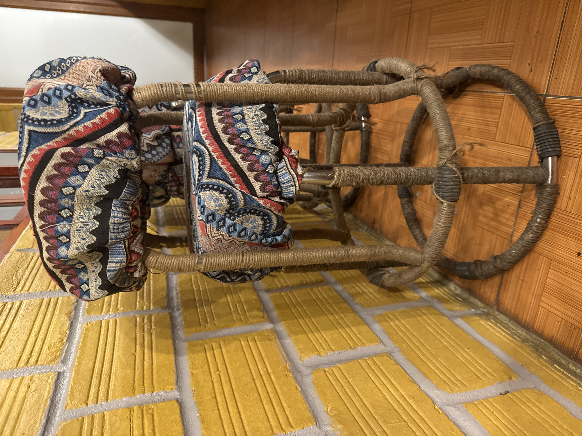 Two rattan stools with fabric cushions are partially visible. The foreground is a faux-brick tiled floor, with walls covered in orange tile. The scene is likely located within a building in or near Mèo Vạc Commune, Vietnam.