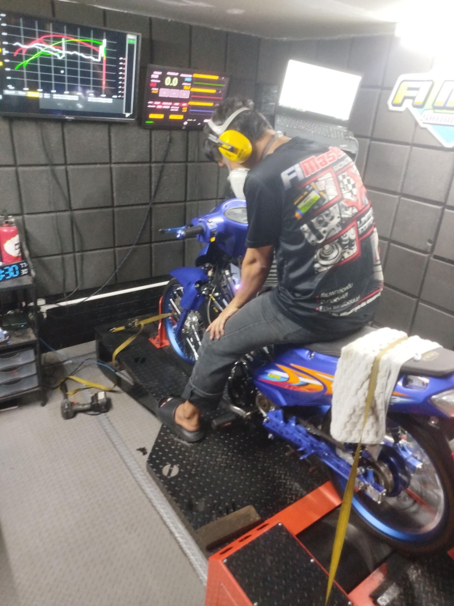 In Ban Kho En, Thailand, a person wearing a black t-shirt with graphics and yellow headphones sits on a blue motorcycle undergoing testing on a dynamometer. The motorcycle is secured to the dynamometer with straps. Two screens are visible on the wall behind the person: one displaying a graph and the other showing numerical data. A laptop is also present on a shelf. The environment appears to be a workshop or testing facility, indicated by the sound-dampening wall panels and various equipment.
