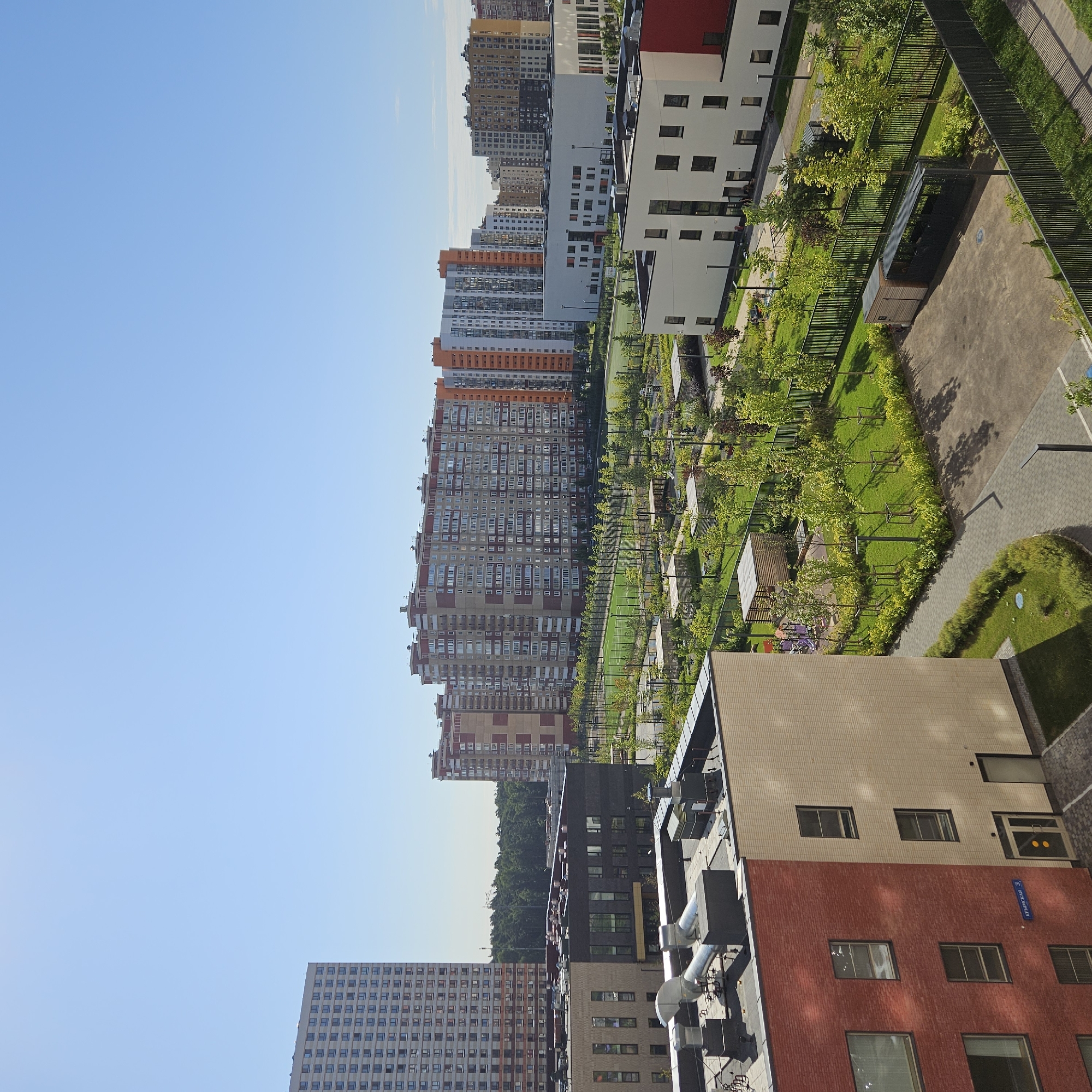 This is an aerial, wide-angle photograph of a modern residential development on a clear, sunny day. In the foreground, a red and beige brick building is partially visible, with several windows and a doorway. Above this, on the roof, are various mechanical units and ventilation pipes.
Behind this building, a complex of low-rise, dark gray and tan buildings forms a courtyard area. This courtyard features manicured lawns, trees, paved walkways, and outdoor recreational spaces including a playground with colorful equipment and wooden gazebos. A black metal fence encloses this area.
In the middle ground, a series of taller apartment buildings are visible. These buildings vary in color, with some featuring red, beige, and orange facades. A sports field, likely a soccer or basketball court, is located between these buildings and a dense green forest in the background.
Further in the distance, more apartment buildings of varying heights and architectural styles are visible, some with yellow and gray exteriors. The sky is a clear, bright blue with no clouds. The image appears to depict a new or recently developed urban neighborhood with a focus on green spaces and amenities for residents. Based on the street sign that reads "Крымская 3" (Krymskaya 3) on the red brick building, this location is in the city of Bobrovo, Russia. There are no people visible in the frame.