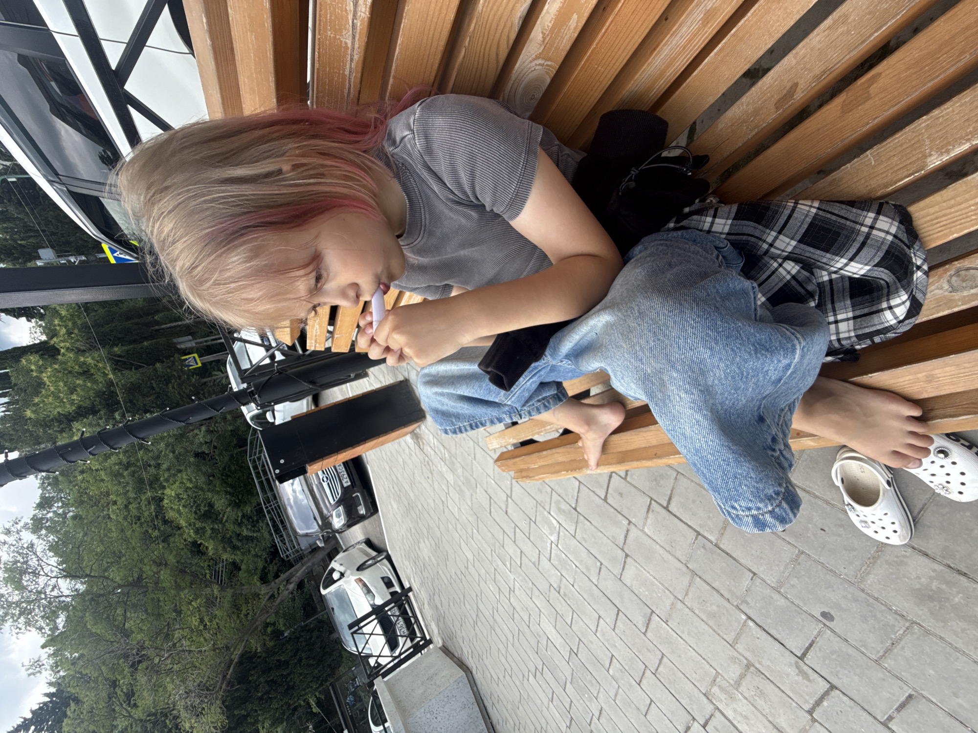 A young blonde girl with pink-dyed hair sits on a wooden bench in an outdoor, urban setting. She is wearing a grey t-shirt and blue jeans, with a plaid shirt and black jacket draped over her lap. She holds a white object to her mouth, possibly a straw or a similar item. Two white Croc-style shoes are on the ground beside her feet. The background features trees, a sidewalk, and parked cars. The location is identified as Unknown, Unknown.