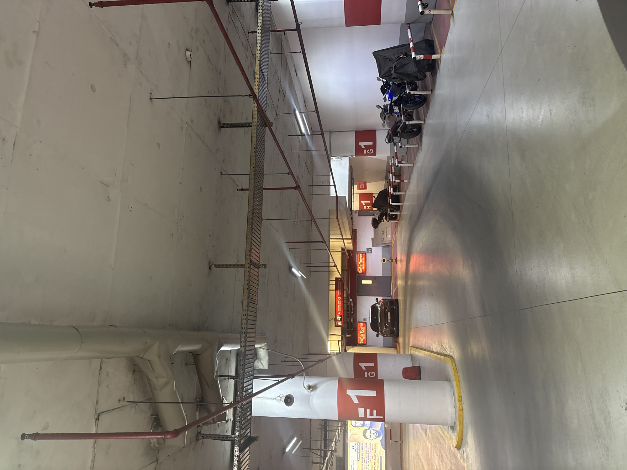 The image shows an indoor parking garage. In the foreground, there are concrete pillars with "F1" and "G1" markings. There are several motorcycles parked in the middle ground, some covered with tarps. A black car is parked further in the background. The ceiling is made of concrete with exposed pipes and electrical conduits. The floor is concrete with yellow lines marking parking spaces. Signs are visible in the background indicating "Sofia Tower" and parking level information. The lighting is artificial, typical of an underground parking facility. The overall scene depicts a functional, albeit somewhat utilitarian, parking area.