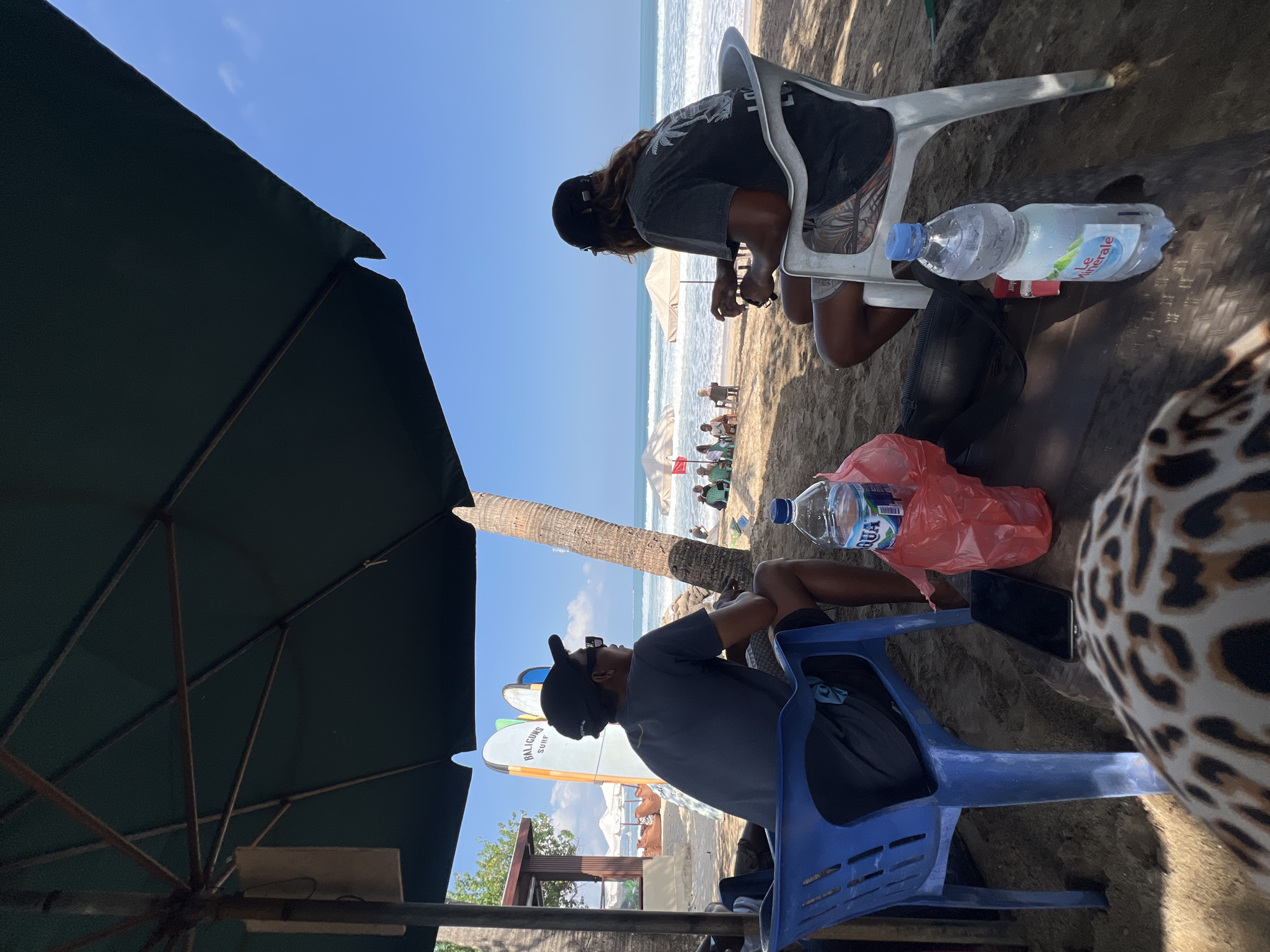 The image depicts a beach scene in Kuta, Indonesia. Two men are seated in chairs under a large green umbrella, facing the ocean. The man on the left wears a dark t-shirt, dark shorts, a black baseball cap, and sunglasses. He is seated in a blue plastic chair. The man on the right wears a dark t-shirt with a palm tree graphic, patterned shorts, and a black baseball cap. He is seated in a white plastic chair. In front of them, on the sandy ground, are two bottles of water, a red plastic bag, a black fanny pack, a smartphone, and a cigarette pack. Surfboards are visible in the background, leaning against a structure. Further in the distance, several beach umbrellas and people are present, and waves are breaking on the shore. The sky is clear and blue.