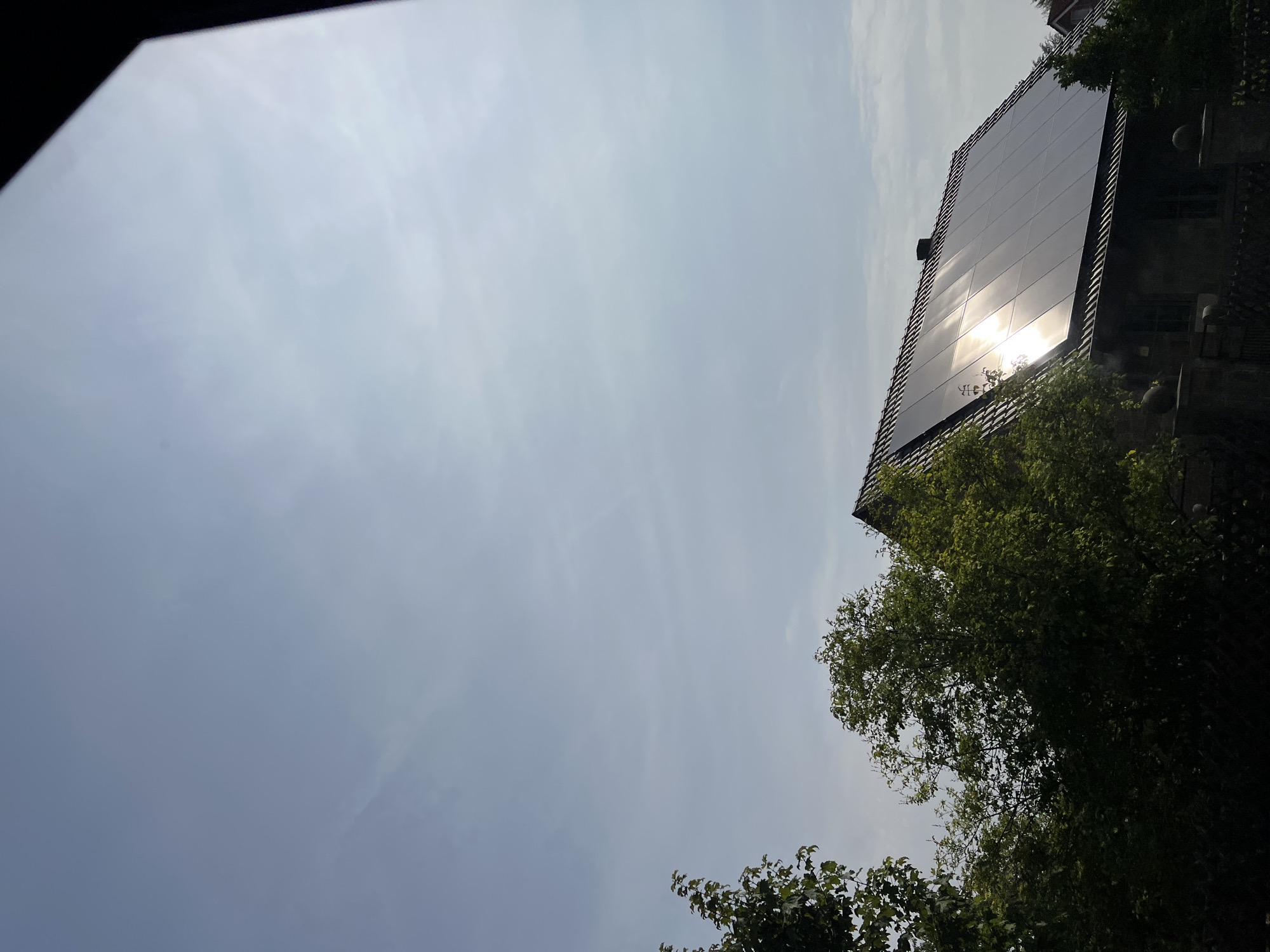 An eye-level, medium shot captures a residential building under a clear sky with some wispy clouds, illuminated by bright sunlight that reflects off the solar panels on its roof. The house, with a dark tiled roof, features solar panels arranged in a grid. Lush green trees and foliage partially obscure the view of the house from the left and center, with a dark fence in the foreground. The scene is set in a suburban environment, indicating a sunny day.