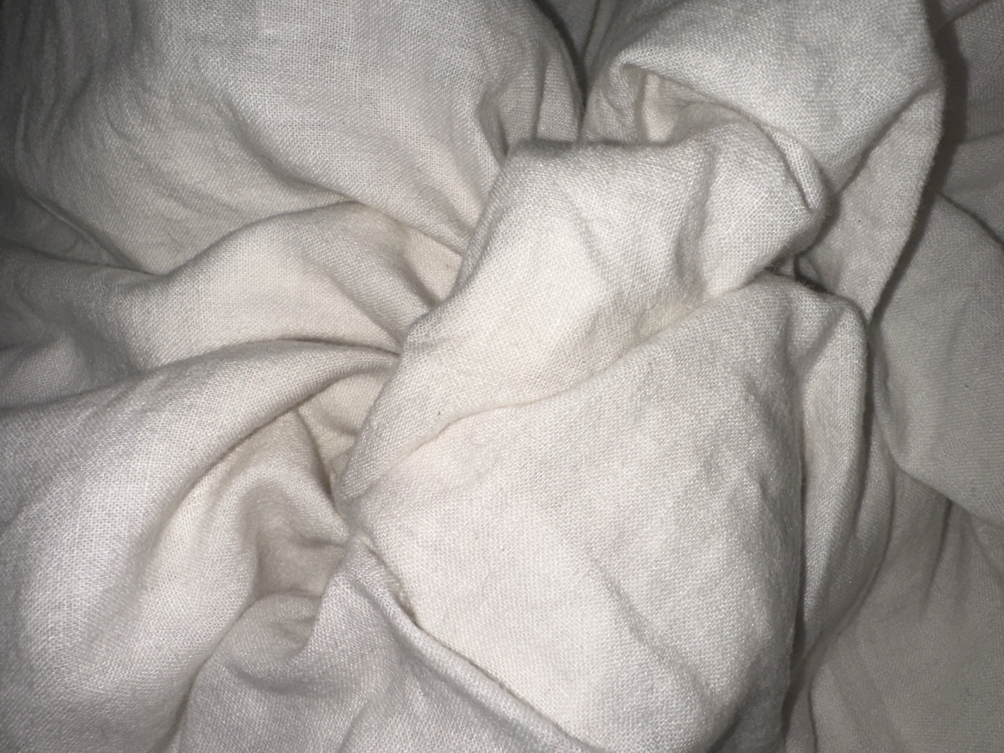The image displays a close-up, top-down perspective of extensively folded and creased light-colored textile, specifically appearing off-white or cream. The fabric exhibits a woven texture with visible individual threads, consistent with a natural fiber such as cotton or linen. Numerous soft undulations, deep folds, and irregular wrinkles create varying patterns of light and shadow across the entire frame. No distinct patterns, foreign objects, individuals, or environmental elements are visible other than the fabric itself. The scene is located in the city of Wealden, United Kingdom.