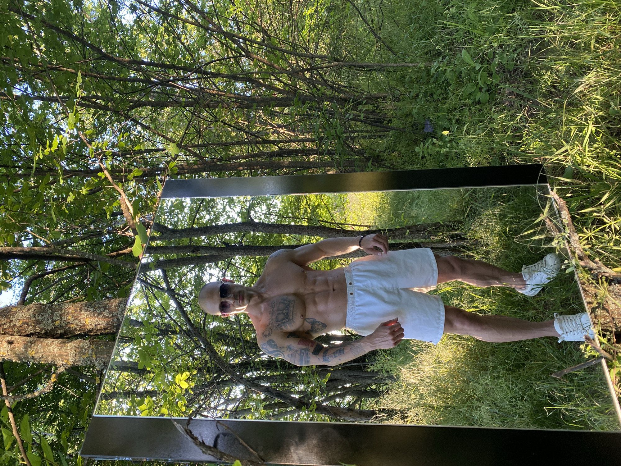 A shirtless man stands next to a large, rectangular mirror placed outdoors in a grassy, wooded area. The man, wearing white shorts and white sneakers, faces the mirror with his bare chest exposed, revealing tattoos on his torso and arms. He also wears sunglasses. The mirror reflects the surrounding trees and sky, creating a layered visual effect. The location is Pleven, Bulgaria, as implied in the request. The environment is characterized by sunlight filtering through the trees and tall green grass.