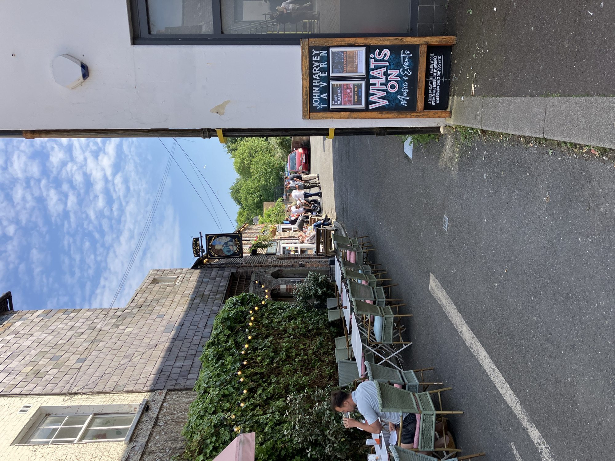 The scene is a street-level view of John Harvey Tavern in Lewes, United Kingdom. The view is framed by an archway. An outdoor seating area with green chairs and white tables extends along the right side of the street. Some people are seated at the tables. A sign advertising "John Harvey Tavern" and "What's On Music & Events" is affixed to the building on the right side of the street. Further down the street, a group of people stand or sit outside the pub. The sky is partially visible with scattered clouds. A sign with "HARVEYS LEWES" is visible above the pub.