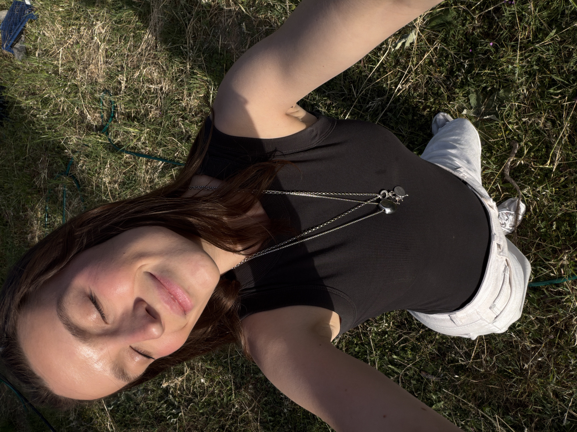 A young adult female is lying on her back in grassy terrain. She is wearing a black ribbed shirt and light-colored denim shorts with grey running shoes. A silver necklace with multiple pendants hangs around her neck. Her eyes are closed, and she appears relaxed. The grass is a mix of green and brown, suggesting a natural outdoor setting. The presence of a blue crate in the background suggests this is a field in Mojanovići, Montenegro.