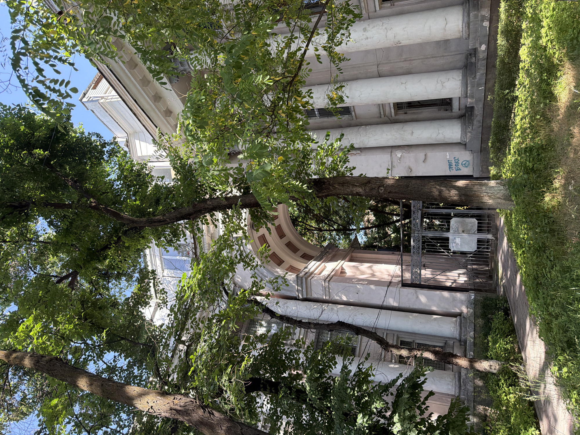 A low-angle view depicts a historic building with prominent pillars and architectural detailing, partially obscured by foliage. The facade shows signs of aging and graffiti. The location is identified as being in Bishkek City, Kyrgyzstan.