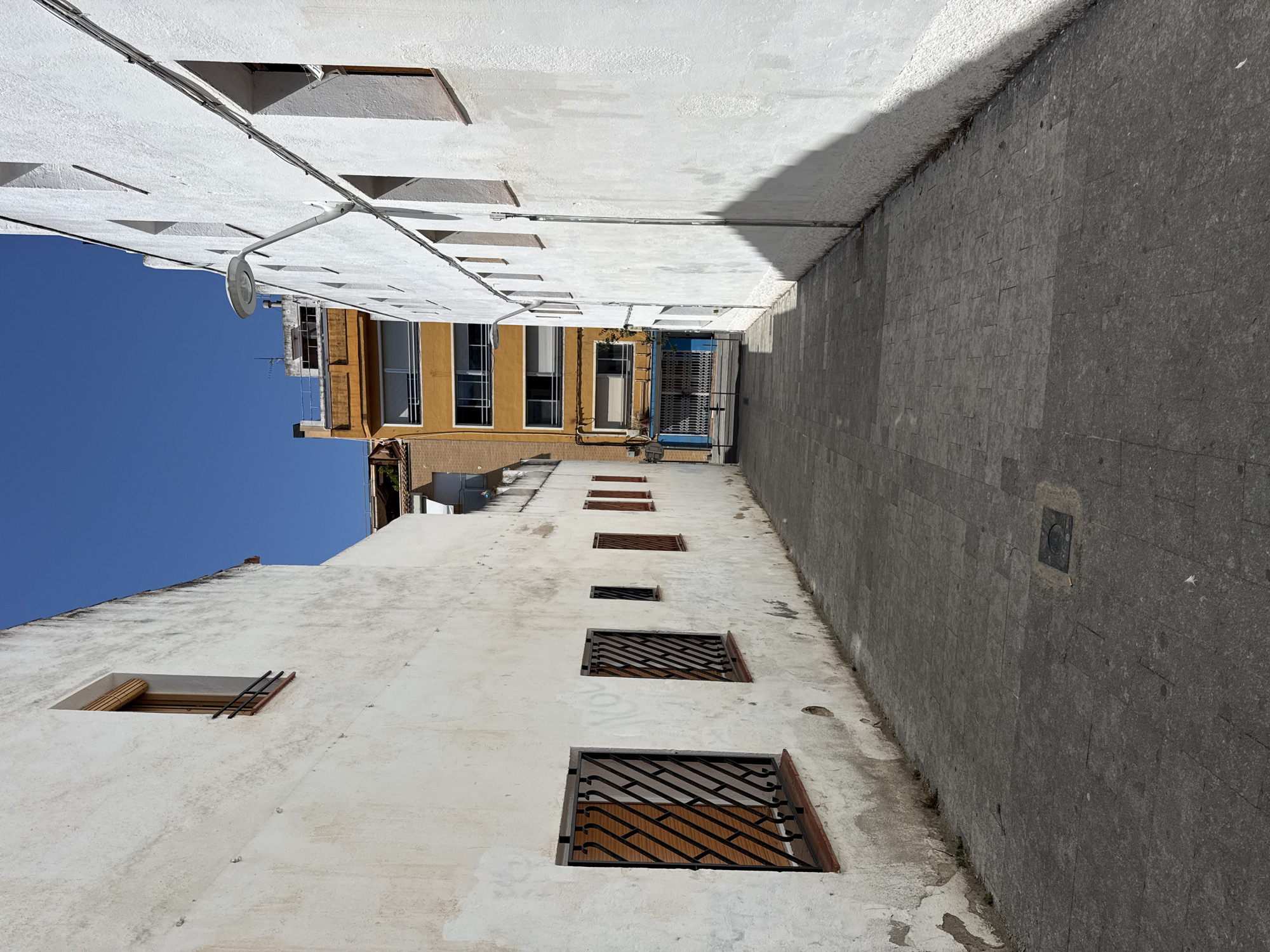 The image depicts a narrow, paved alleyway between two buildings in Blanes, Spain. The building on the left is a two-story structure with a white, textured facade and several windows with dark metal grates. One window on the upper floor has a brown wooden shutter partially lowered. The building on the right also has a white, textured facade and features multiple windows and a white streetlamp mounted on the wall. In the background, a taller, orange building with numerous windows and balconies is visible. The ground is paved with gray interlocking stones, and there is a circular metal grate embedded in the pavement. The sky is clear and blue. No people are visible in the scene.