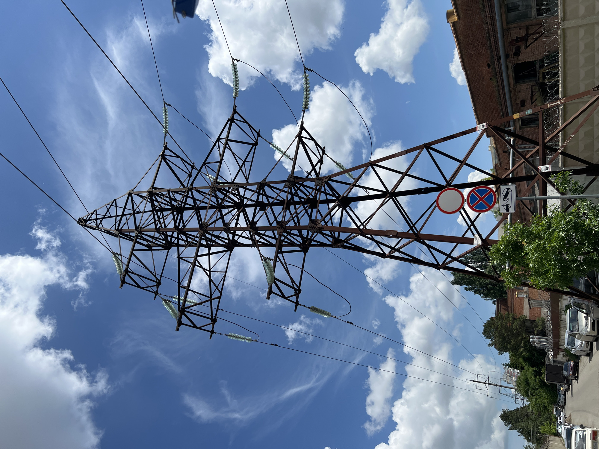 The image shows a steel lattice transmission tower against a partly cloudy blue sky. The tower is situated near a building with a red brick facade and barbed wire along its roofline. The street below features a row of parked cars and green trees. Attached to the tower are several road signs including "No stopping" and "No parking". The image was taken in the city of Krasnodar, Russia.