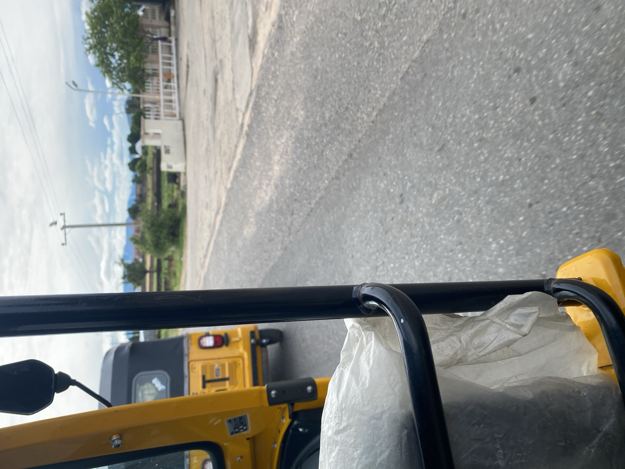This is a first-person perspective, close-up shot taken from inside a yellow auto-rickshaw. The focus is on the interior of the vehicle, specifically a black metal bar across the top of the frame, a white plastic bag filled with what appears to be dark, granular material, and a portion of the yellow body of the auto-rickshaw. A side mirror is also visible on the left.
In the background, the road is visible, and beyond that, a landscape with green trees, some buildings, and a power pole under a cloudy sky with patches of blue. The overall scene suggests a journey in a rural or semi-urban area in Jalingo, Nigeria. The lighting indicates it's daytime, likely mid-morning or afternoon, with overcast conditions. The perspective suggests the auto-rickshaw is in motion. There is no discernible text in the image.