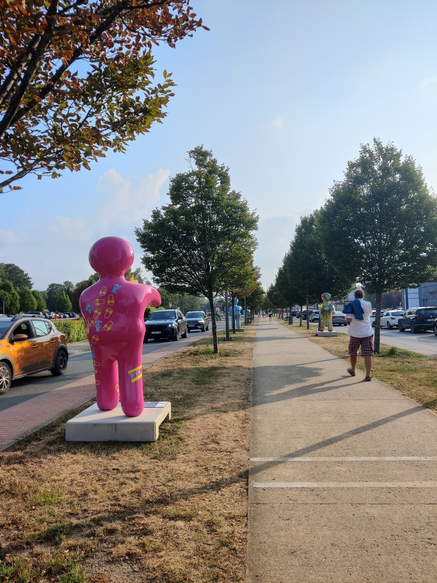 The image depicts an outdoor scene with a sidewalk, a street, and grassy areas. In the foreground, a large, bright pink, anthropomorphic sculpture is positioned on a white base. The sculpture has a smooth, glossy finish and is adorned with various colorful stickers, including some depicting characters and symbols. To the left of the sculpture, an orange car is parked on the street.
Further down the street, there are more similar sculptures, some in blue and green, lining the sidewalk. Several cars are driving on the street, and a few people are walking on the sidewalk. One individual in the mid-ground is walking away from the camera, wearing a white shirt, plaid shorts, and sandals, carrying a blue garment over their shoulder. The environment appears to be a public space, possibly a commercial or entertainment area, with trees and buildings visible in the background.
The scene is bathed in natural sunlight, suggesting it is daytime. The overall impression is one of a public art installation or a pedestrian-friendly area with decorative elements. This image was captured in Marche-en-Famenne, Belgium.