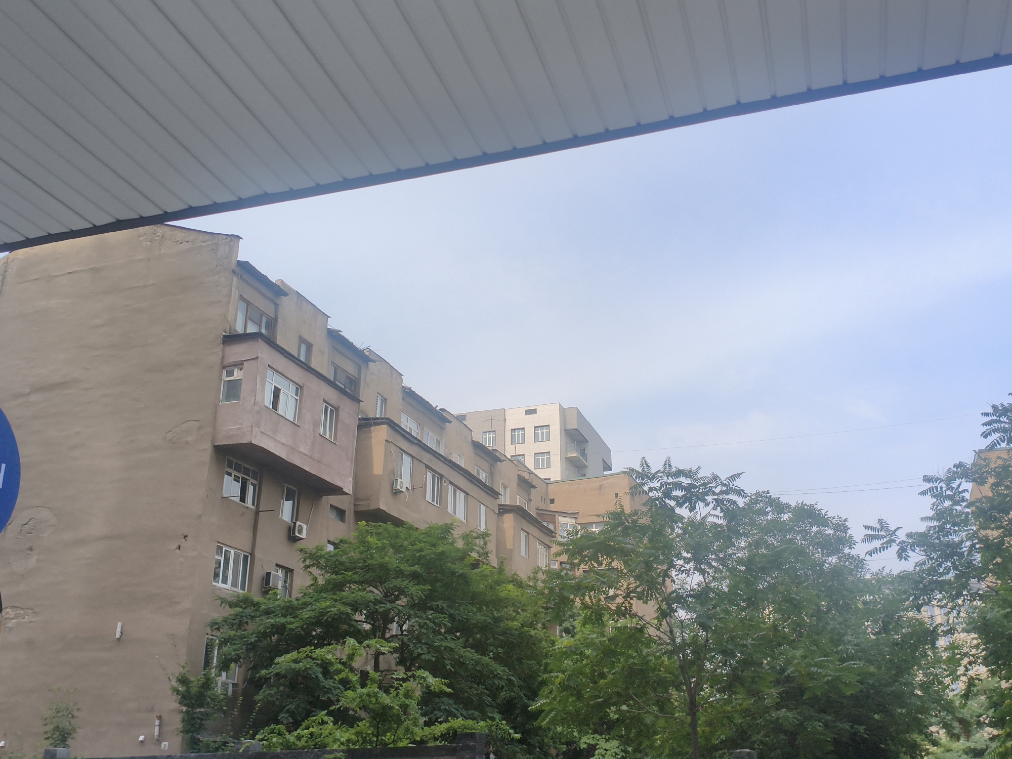 The image depicts a street view in a city, with a prominent multi-story building dominating the left side of the frame. The building has a beige, weathered facade and features numerous windows, some with white frames and others with darker frames. A protruding section with windows juts out from the main structure on the third and fourth floors. Several air conditioning units are visible on the exterior walls. Trees with lush green foliage are in the foreground and midground, partially obscuring the lower levels of the building. In the background, another modern-looking building with a lighter facade and a grid of windows is visible. The sky is a pale blue with a few wispy clouds. The top of the frame is partially covered by a metallic, ribbed overhang, likely part of a structure in the foreground. A partially visible blue circular sign with white markings is on the far left of the building. The scene appears to be in an urban environment under a cloudy sky.