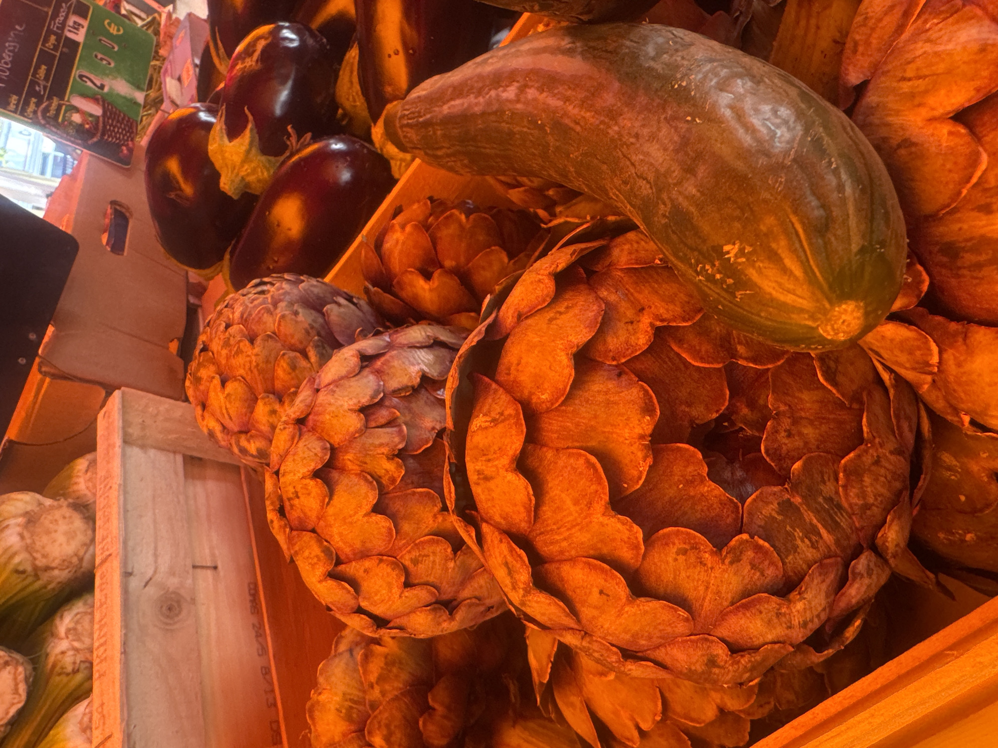 In Saint-Claude, France, at a market stall, a close-up shot captures a variety of fresh produce. Dominating the foreground are several large artichokes, their outer leaves a warm, earthy orange hue, revealing intricate layers. To the right, a long, dark green zucchini lies diagonally across the artichokes. In the background, more artichokes are visible, alongside glossy, dark purple eggplants. A wooden crate with a visible label and some loose celery stalks add to the market scene. The lighting casts a warm, golden glow over the entire arrangement.