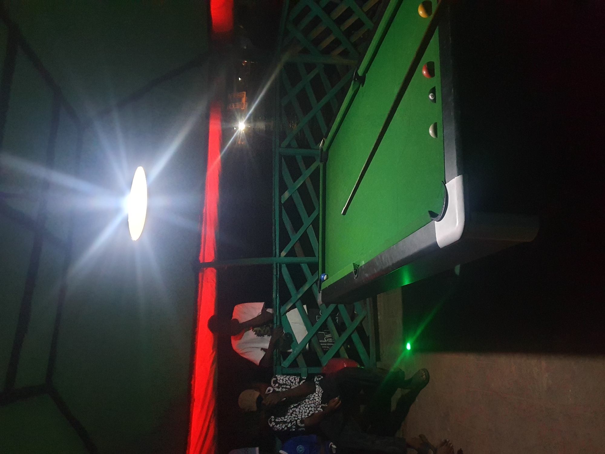 The image depicts an outdoor pool hall with a green canopy overhead, illuminated by a bright overhead light that causes lens flare. Red fabric is visible along the edge of the canopy. A green wooden railing separates the pool area from the background, which is dark and appears to be a body of water.
In the foreground, a green pool table is set up, with several billiard balls visible on its surface, including a white cue ball, a red ball, an 8-ball, and a yellow ball. A pool cue rests on the table.
To the left of the pool table, three individuals are present. One person, wearing a patterned shirt and a light-colored cap, is seated on a chair. Another person, wearing a white t-shirt with a graphic design, stands near the railing, facing away from the camera and resting their chin on their hand. A third person, wearing a blue shirt, is partially visible on the far left.
Green laser lights are visible on the floor in the lower left and lower right sections of the image. The scene suggests a casual gathering or recreational activity in Lokoja, Nigeria.