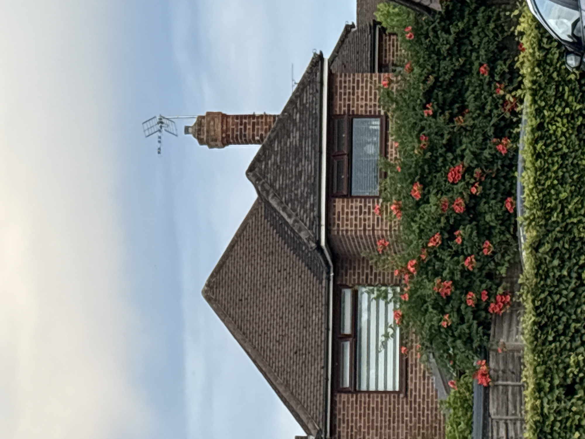 The image shows the side of a brick house in Chelmsford, United Kingdom, with a prominent roof and chimney. The house features two windows, one on the left with horizontal blinds and another on the right. A large bush with red flowers is situated in front of the house, obscuring a wooden fence. The sky above is partly cloudy.