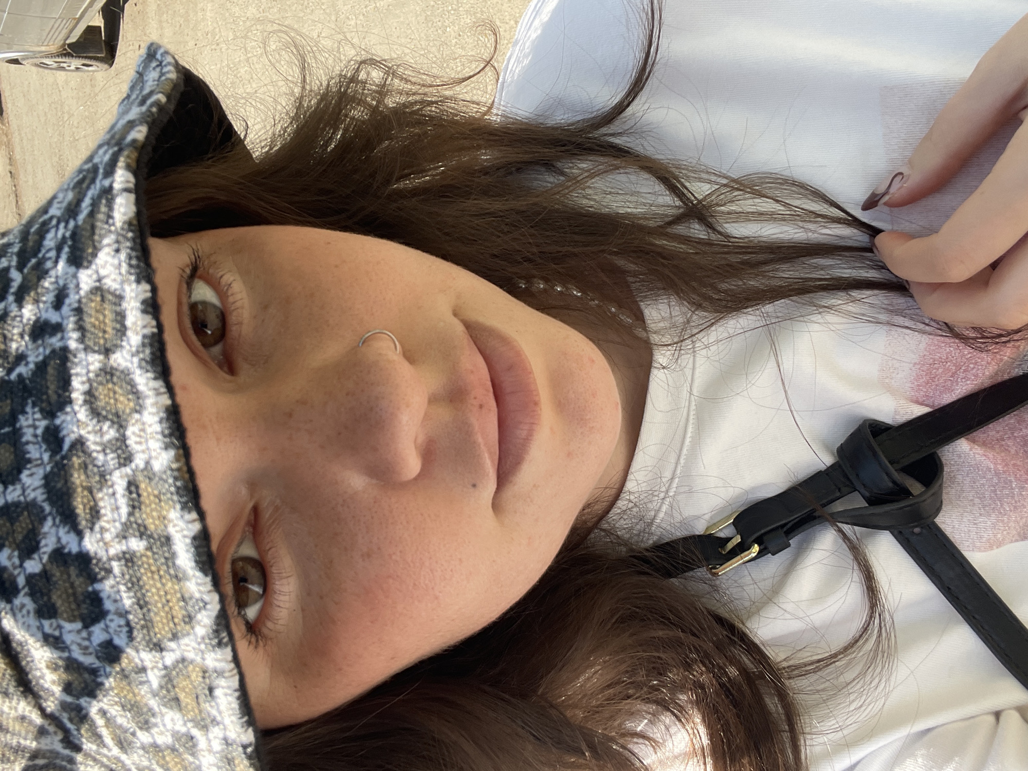 A person is lying down, face towards the camera. The individual has fair skin, brown hair, and brown eyes, with a nose ring on the left side. She is wearing a patterned hat and a white shirt. A black leather strap with a knot is partially visible over the shirt. The person's right hand is holding a strand of her own hair. The background appears to be an outdoor concrete surface. There is no clear evidence to indicate the location is in Izhevsk, Russia.