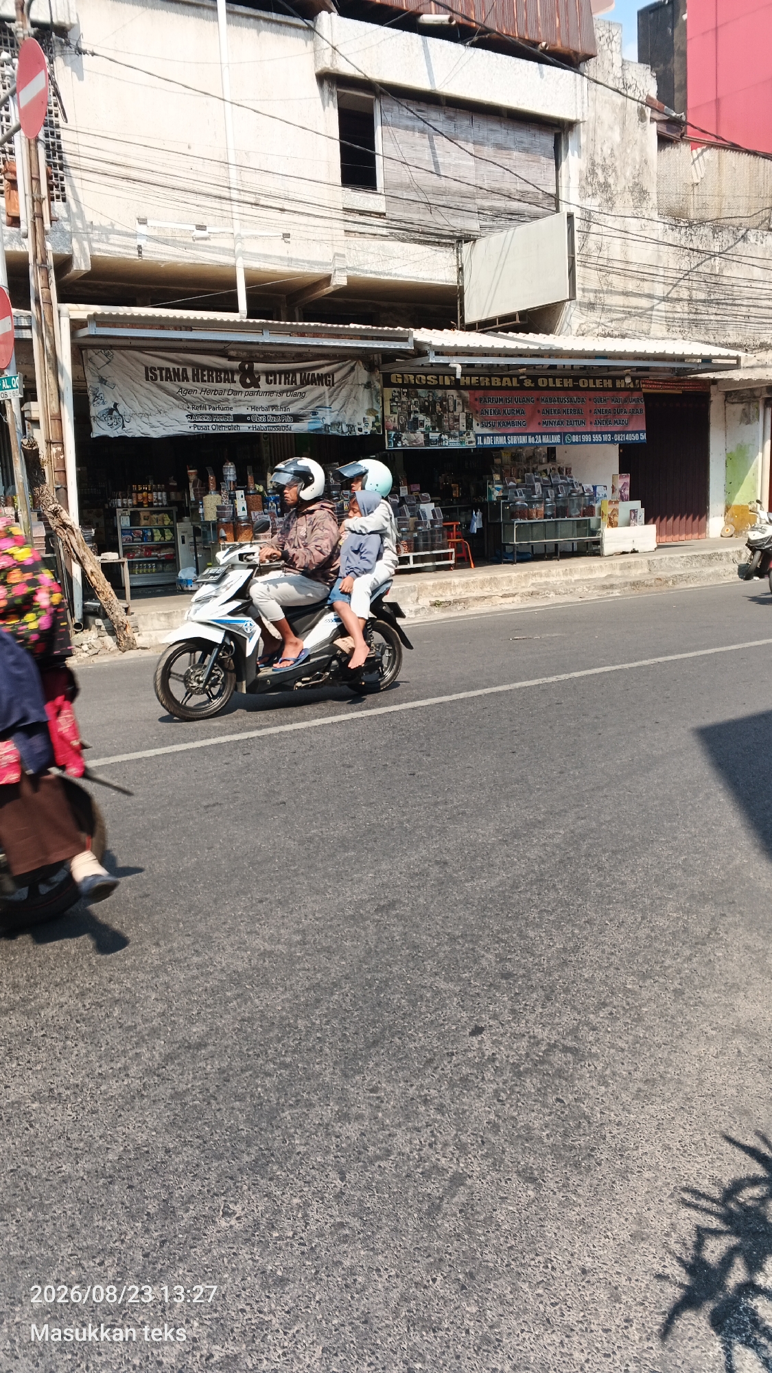 A medium-wide shot captures a sunlit street scene in Malang, Indonesia. A man wearing a patterned jacket and white pants drives a white scooter with two children. The man has a white helmet, and the child behind him wears a light blue helmet and jacket. The child in front wears a blue hoodie. They are traveling on a paved road with visible lane markings. To the left of the road, a woman with a colorful patterned top and brown skirt is partially visible on another scooter, her leg and foot in view. Behind them, storefronts line the street, displaying goods and signage. One banner reads "ISTANA HERBAL & CITRA WANGI". Another sign indicates "GROSIR HERBAL & OLEH-OLEH MALANG". The buildings are light-colored, and the sky is bright and clear, suggesting a warm day. A timestamp at the bottom reads "2026/08/23 13:27".
