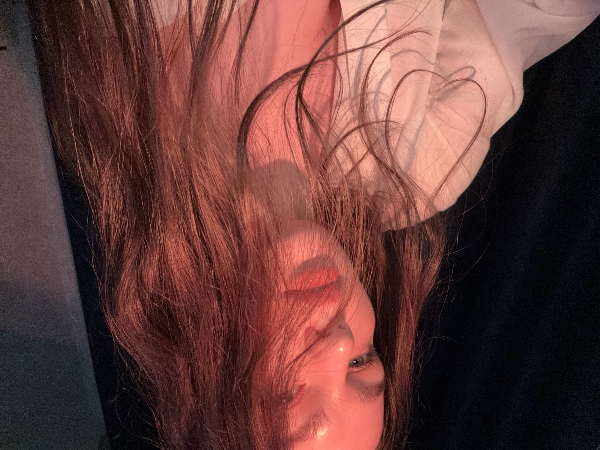 A person with long brown hair is lying down, possibly on a couch. The person is wearing a light-colored shirt. The location of the image is not discernible from the visual information.