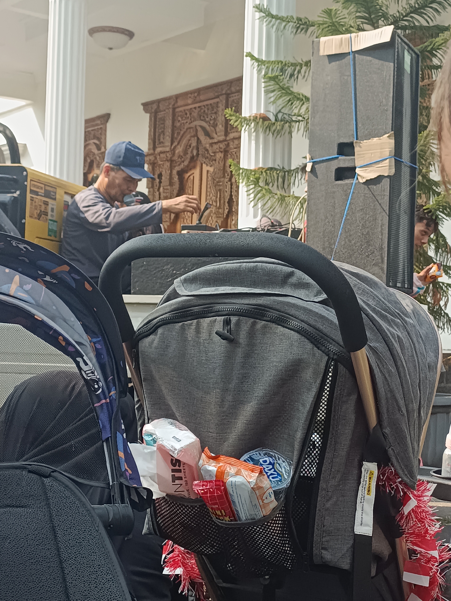 In Sleman, Indonesia, a man wearing a blue baseball cap and a grey long-sleeved shirt is speaking into a microphone while operating equipment. He is standing behind a stroller. In front of the stroller, there are packages of tissues, snacks, and a round blue object, possibly a disc. To the right of the man, a large black speaker is tied with blue rope and secured with cardboard. A child is visible behind the speaker, looking towards the stroller. Another stroller, partly visible on the left, is patterned with abstract shapes and cartoon characters. The scene appears to be set outdoors, with a pine tree and decorative carved wooden panels in the background.