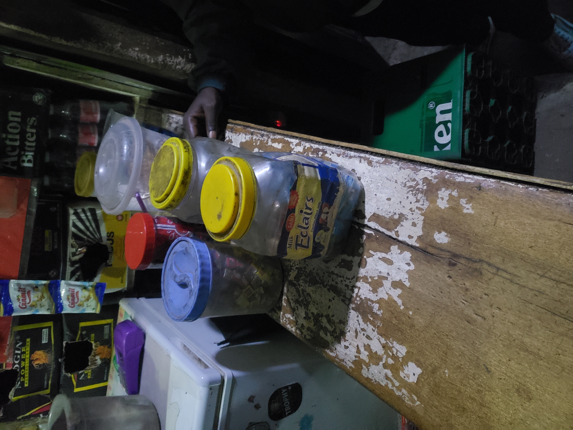 The image depicts a dimly lit interior, likely a small shop or kiosk, with a focus on various food items and beverages. In the foreground, a wooden counter shows signs of wear and peeling paint, with several glass jars containing food products. One jar is labeled "Milk SANI Eclairs," suggesting it holds candy or similar confectionery. Next to it, a plastic container with a blue lid and another with a red lid are visible, also appearing to hold food items. Behind these, a larger, clear plastic container with a yellow lid is partially obscured.
To the left, a white appliance, possibly a small refrigerator or cooler, is present with a sticker that reads "TROPHY." A purple object, resembling a small electronic device or accessory, sits on top of the appliance. In the background, shelves are stocked with various items, including what appears to be boxed drinks and bottles of beverages. A prominent sign displays "Action Bitters." The overall impression is of a typical Nigerian local shop.
The scene is consistent with what one might find in a commercial area of Jos, Nigeria, characterized by local businesses selling everyday commodities. The lighting suggests it might be evening or a poorly lit indoor space. The presence of a Heineken crate in the lower right corner further reinforces the commercial setting. No specific individuals are clearly identifiable or engaged in significant actions.