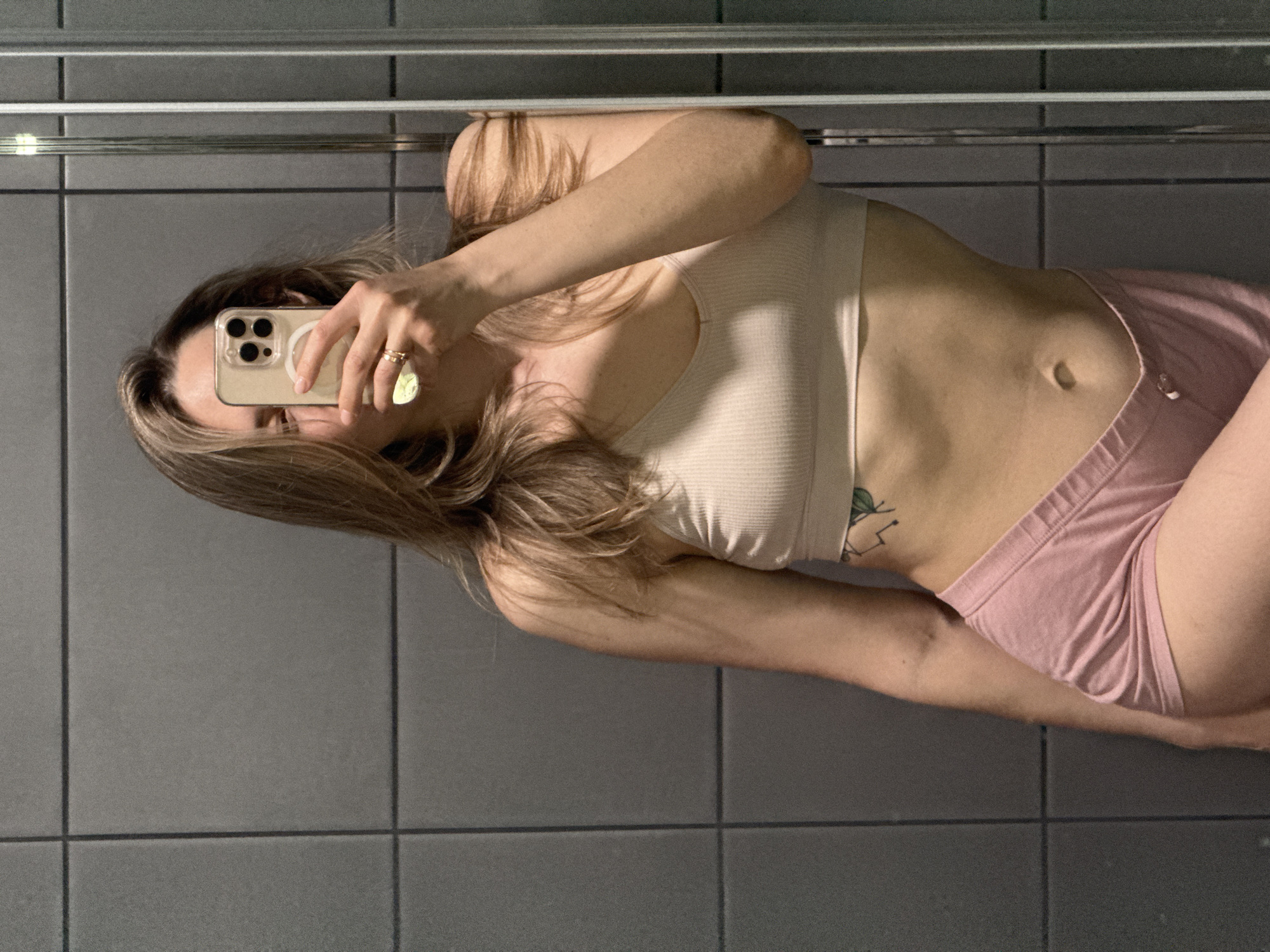 A woman is taking a mirror selfie. She is wearing a white ribbed tank top and pink shorts. The background is a tiled wall. The location cannot be determined from the image.
