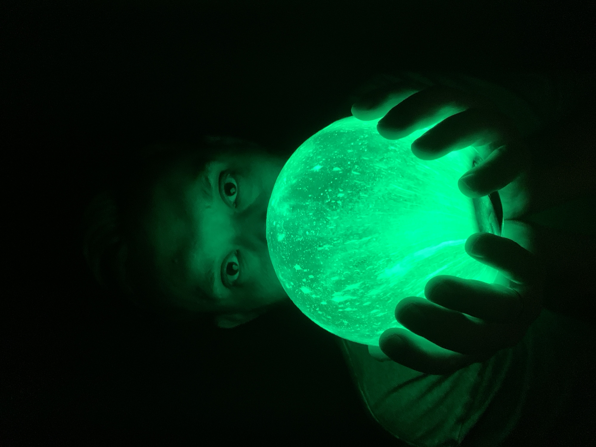 A child's face and hands are illuminated by a glowing green orb they hold. The scene is dark except for the green light emanating from the object, which casts a bright glow on the child's face, hands, and a portion of their shirt. The child's eyes are wide and appear to be looking directly at the viewer. The backdrop is undifferentiated darkness. The city of Chaikovsky, Russia is not identifiable in the image.